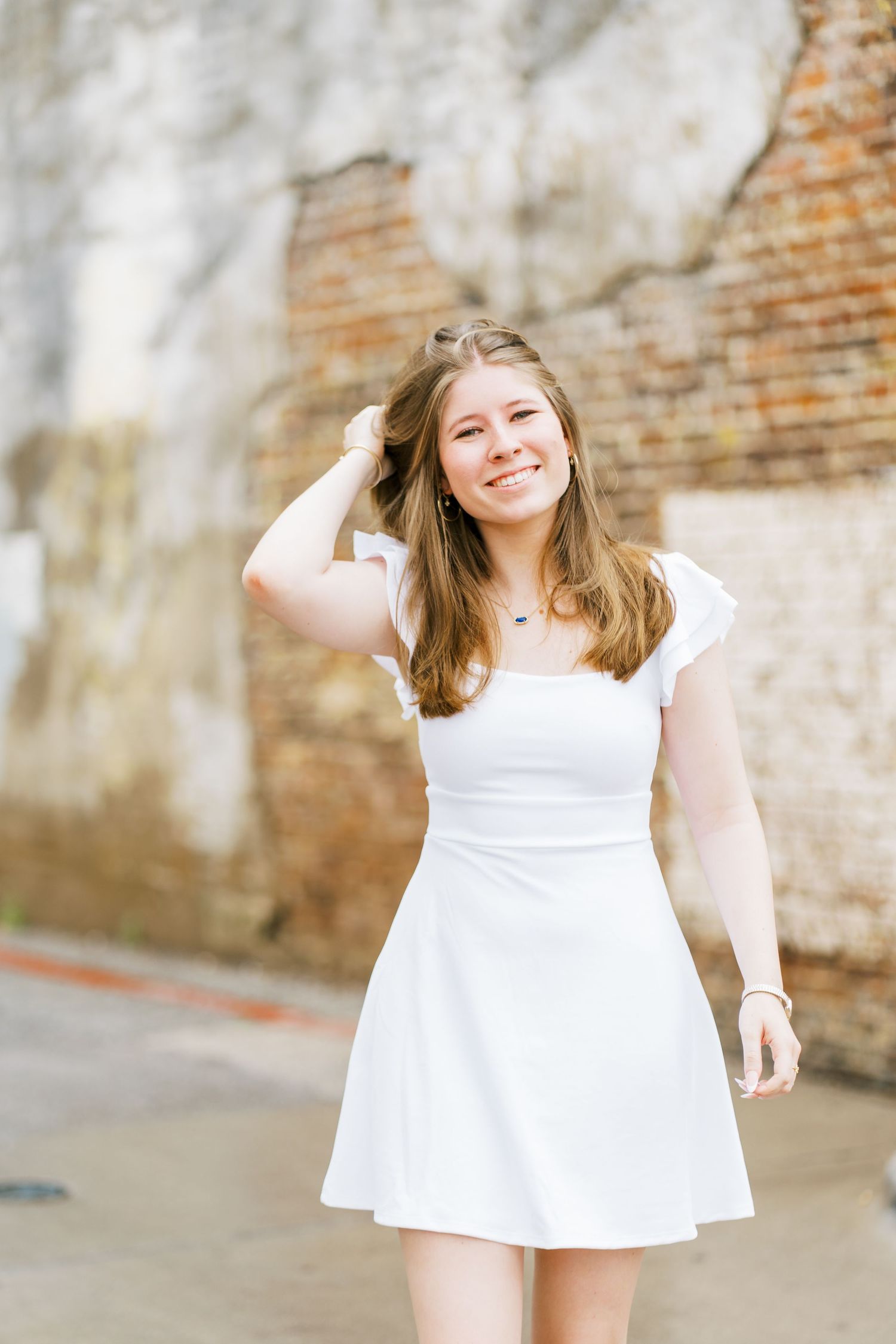 A person in a white summer dress poses casually against a weathered brick wall background.