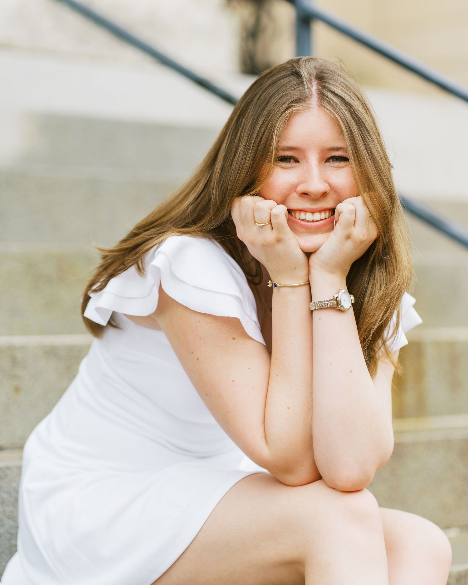 Smiling person in white dress sitting on concrete steps while resting chin on hands in outdoor portrait.