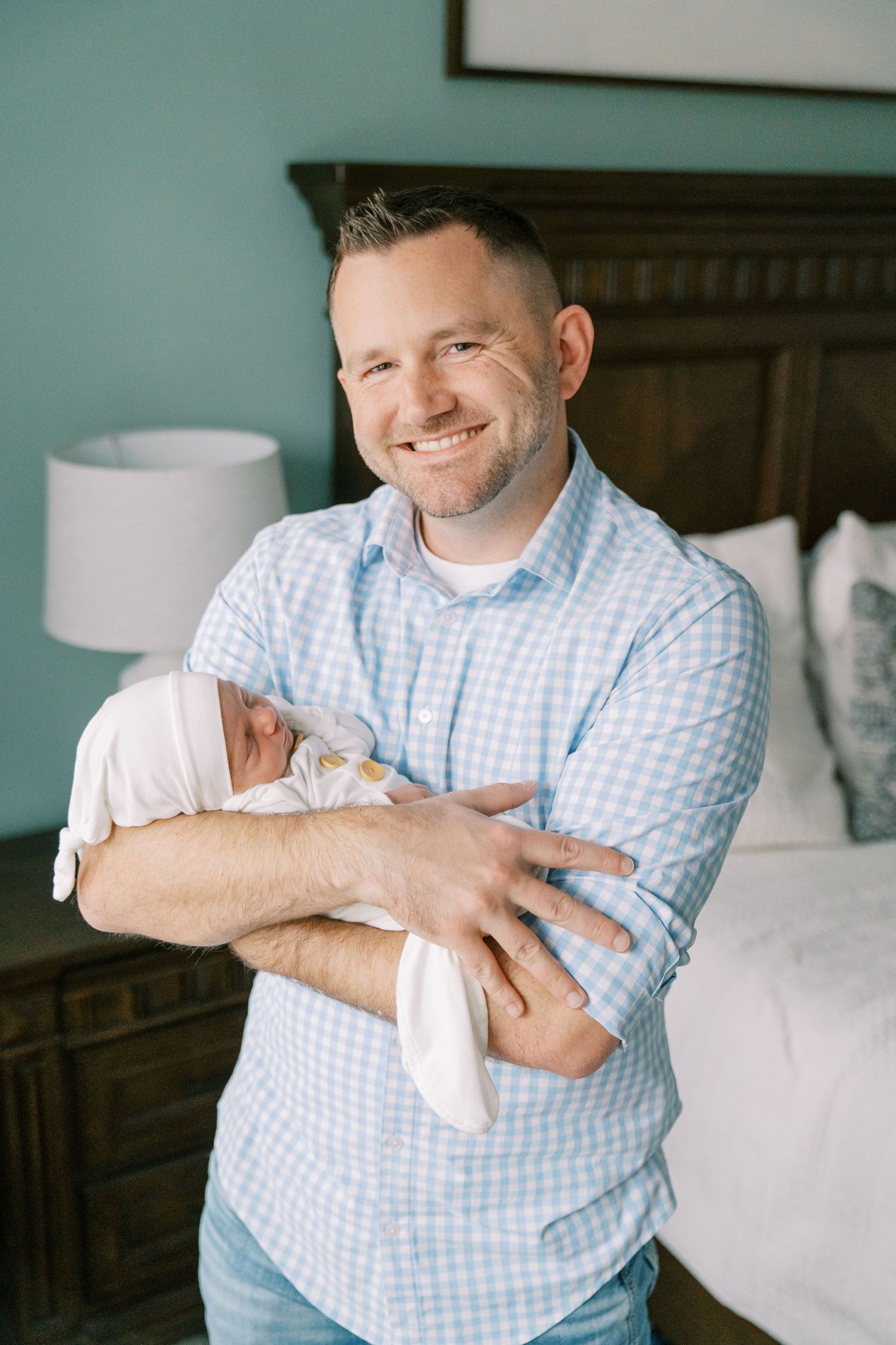 Parent in blue checkered shirt holding and looking lovingly at newborn baby in bedroom setting.