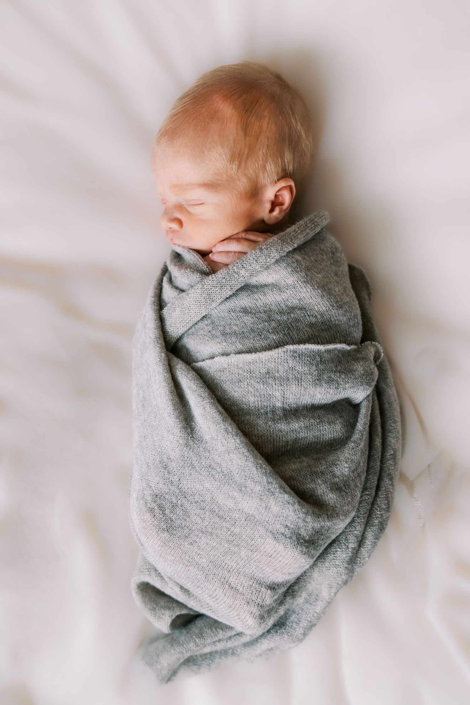 Newborn baby wrapped in soft gray blanket lying on white bedding.