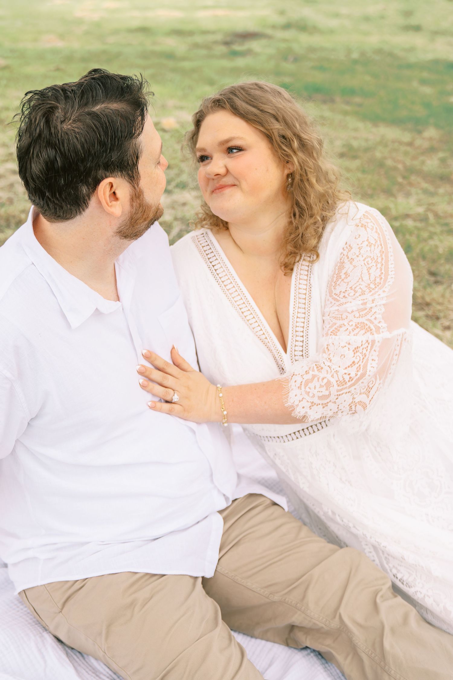 Bride in lace white dress gazing at groom during romantic outdoor engagement photo session.