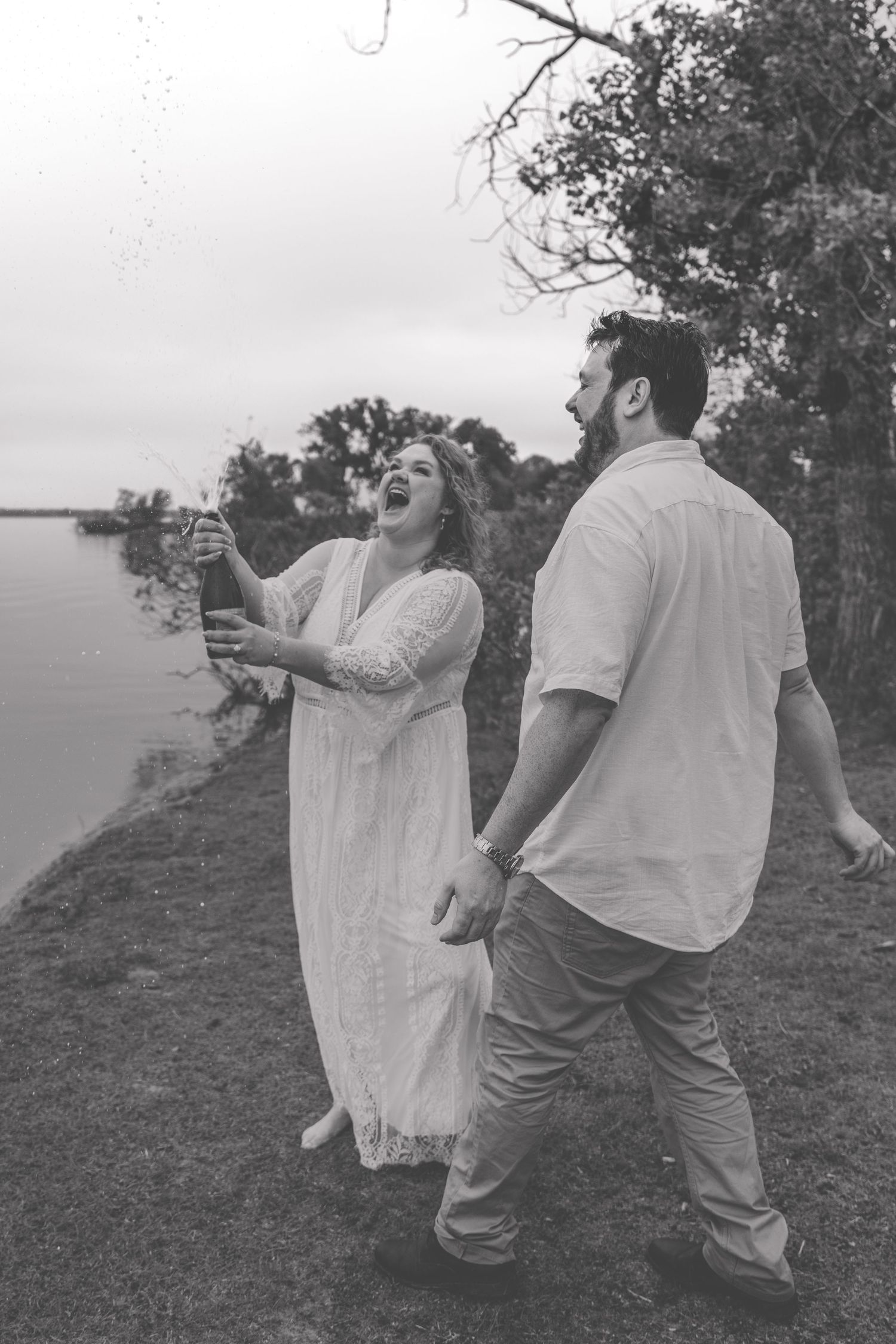 Black and white photo of laughing couple popping champagne bottle near lakeside during celebration.