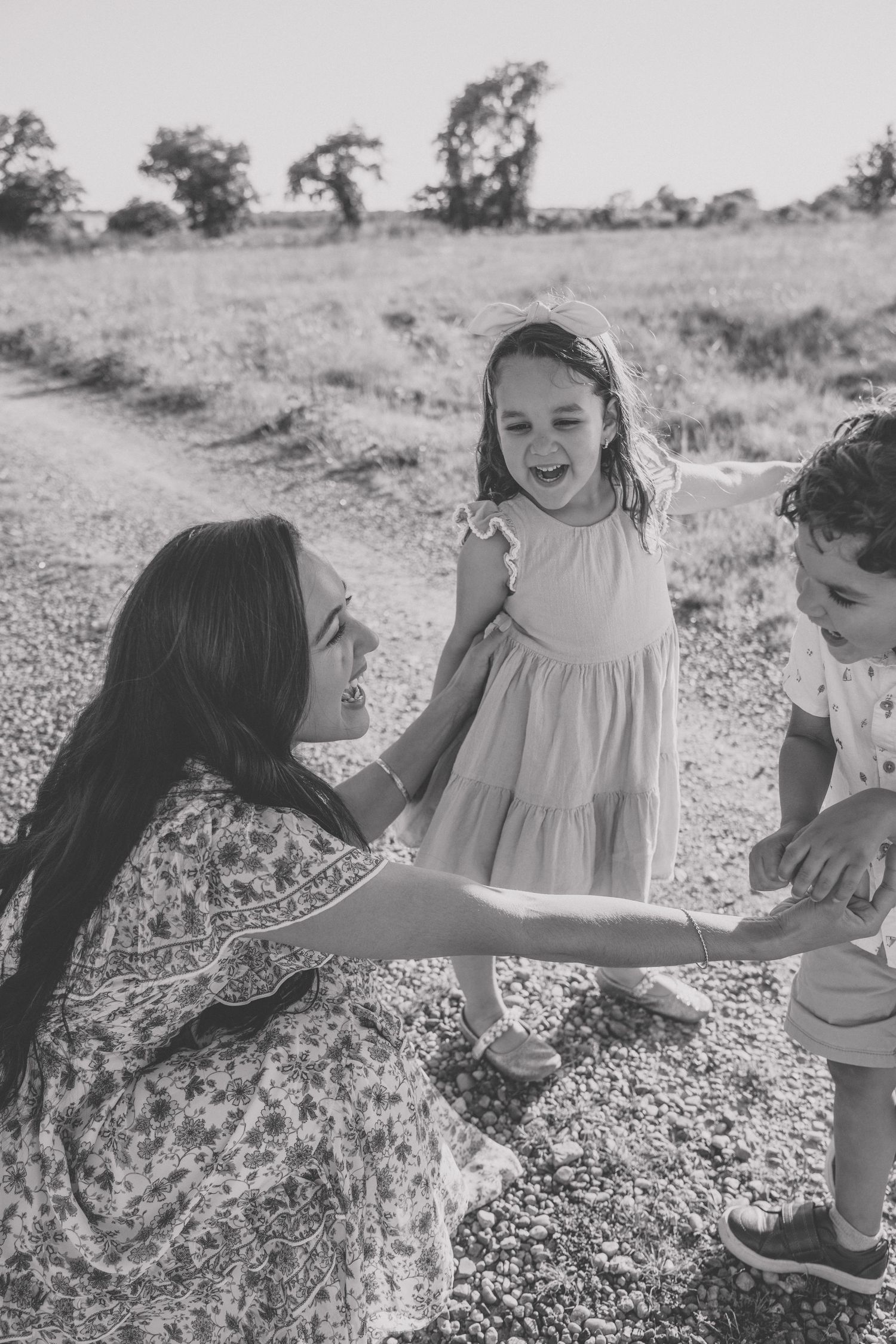 Black and white photo of a mother kneeling with two young children on a rural path, sharing a joyful moment outdoors.