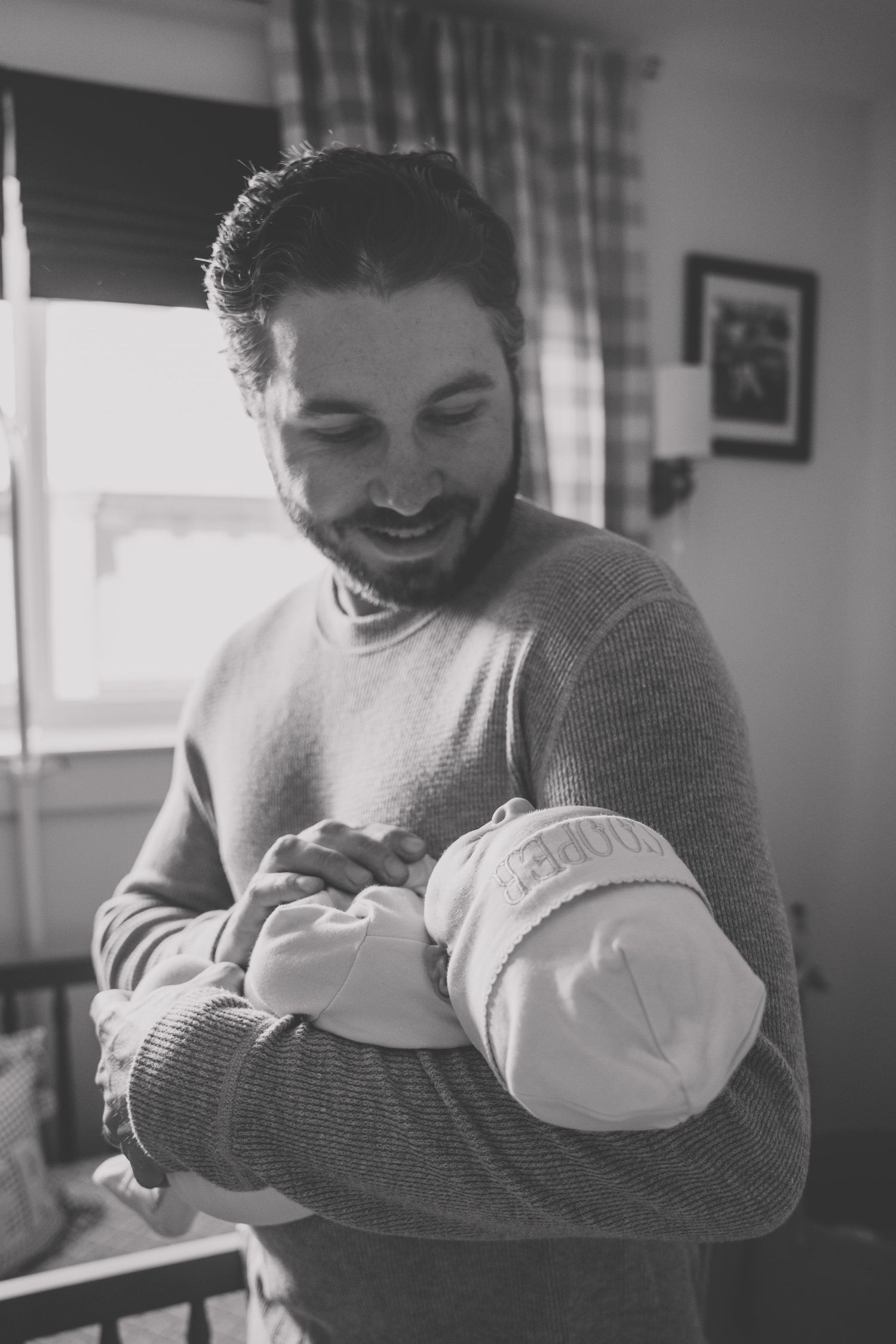 Smiling bearded man cradling a newborn baby in his arms indoors, black and white photo.