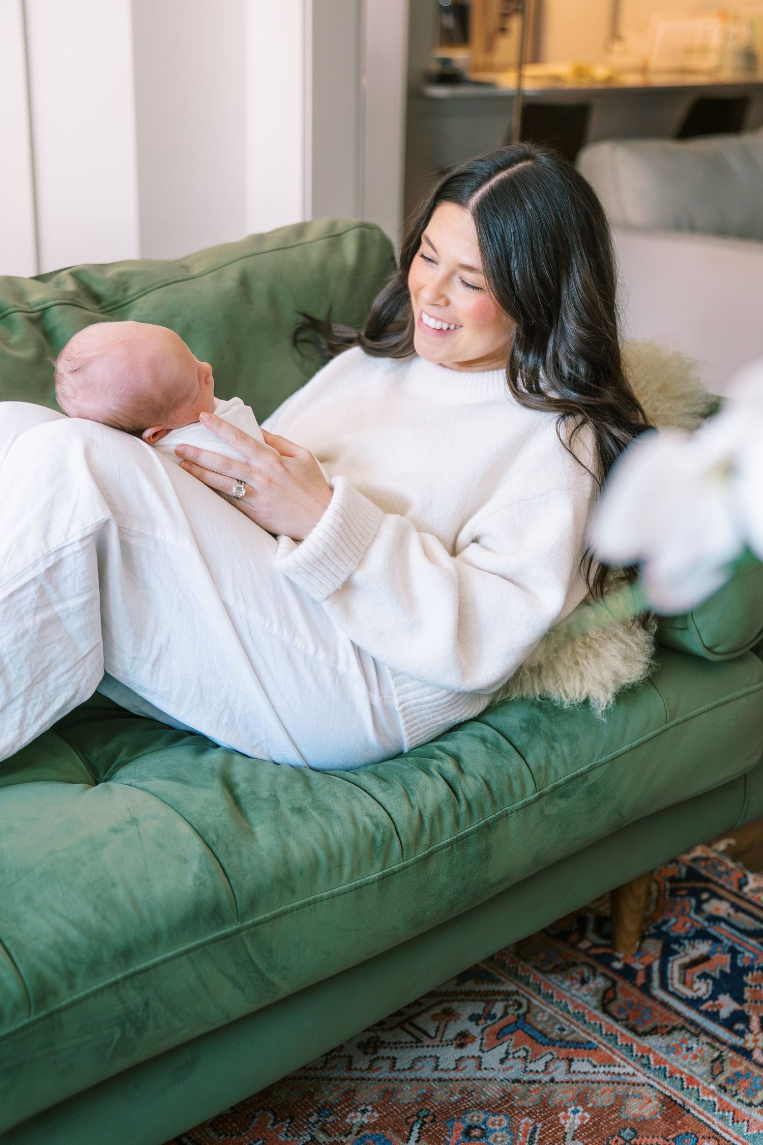 A smiling dark-haired mother in a cream sweater reclines on a green velvet sofa, lovingly holding her newborn baby.