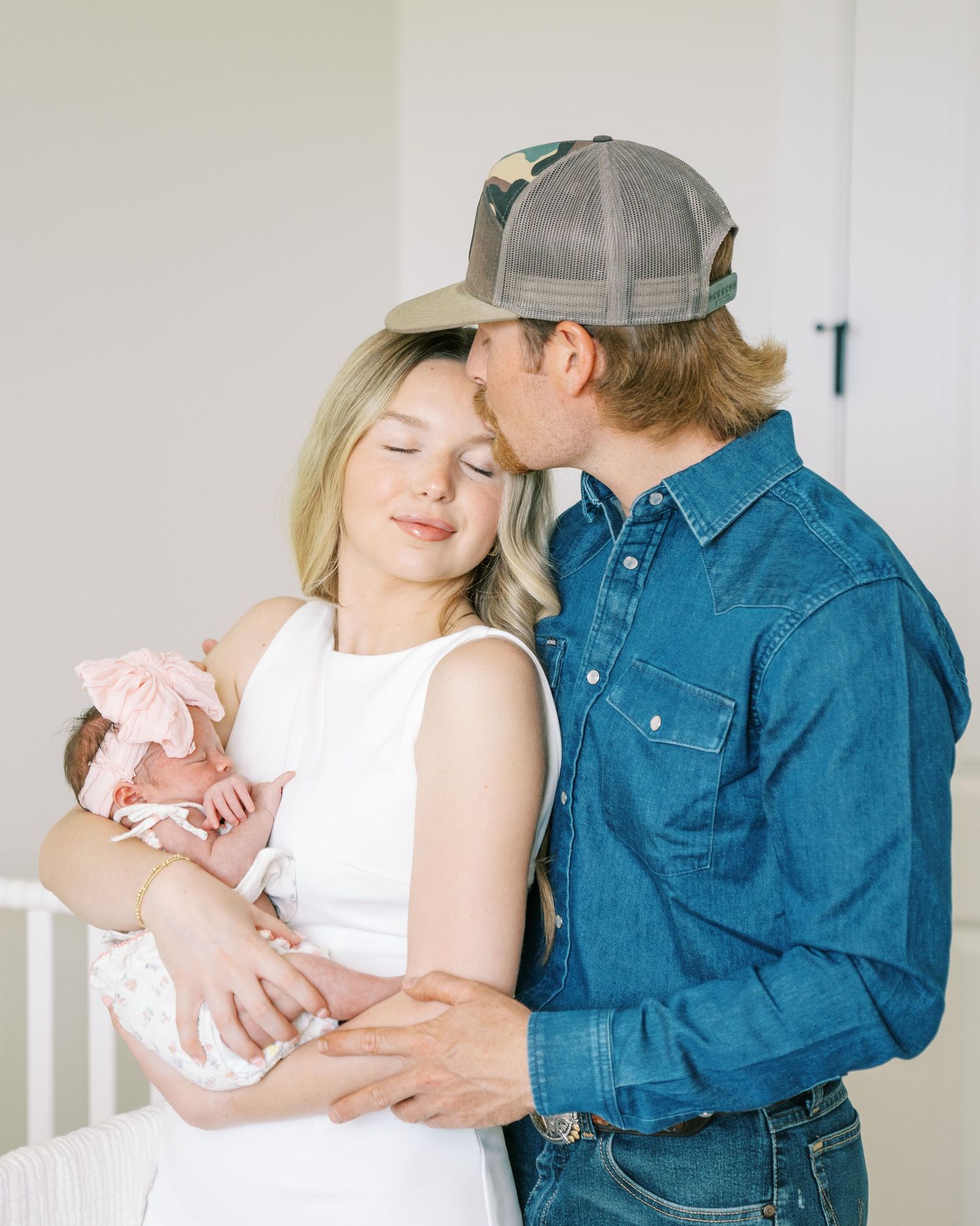 A couple shares a tender moment with their newborn baby, the man kissing the woman's head as she holds the infant.