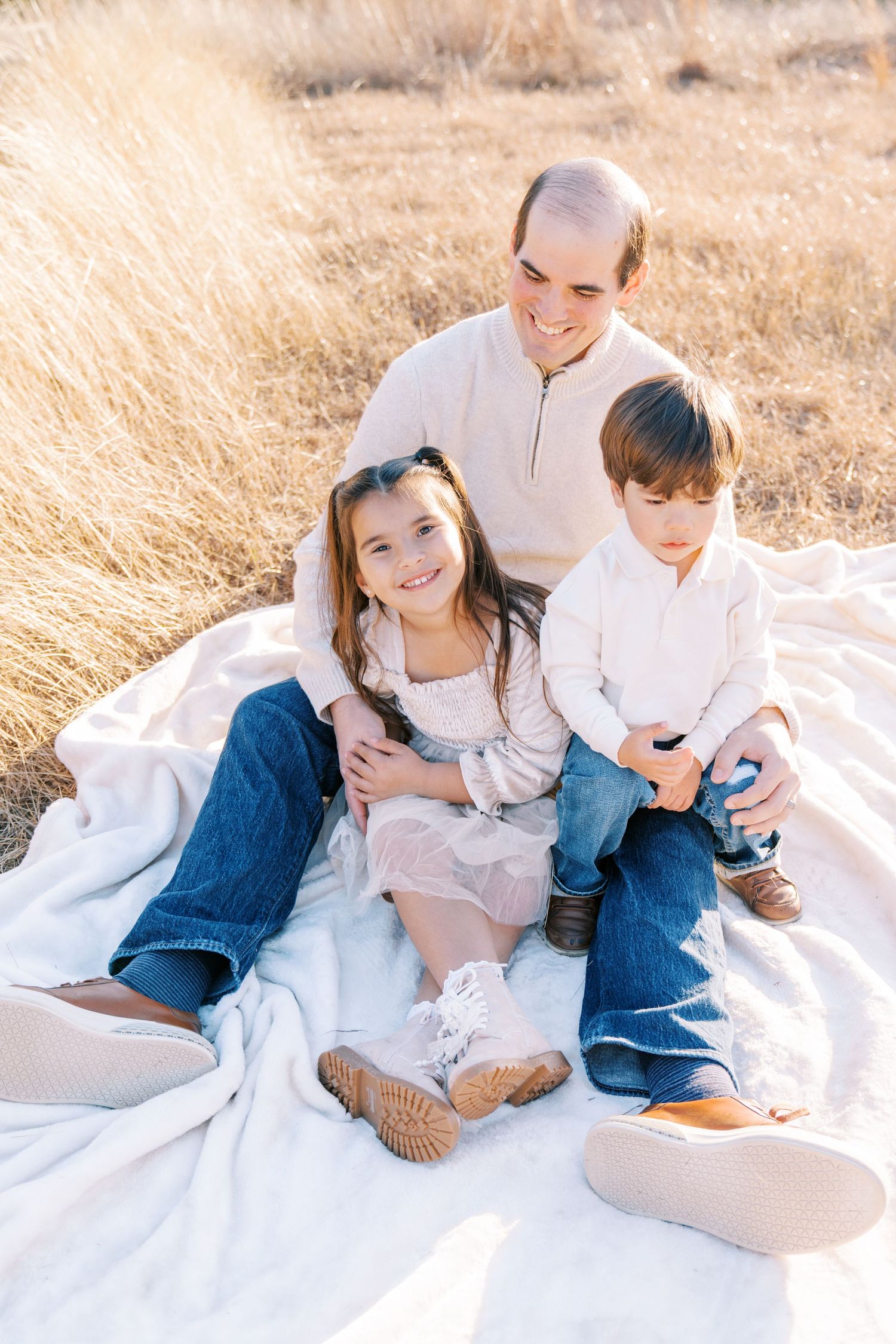 Family relaxing together on white blanket in golden wheat field during sunset.