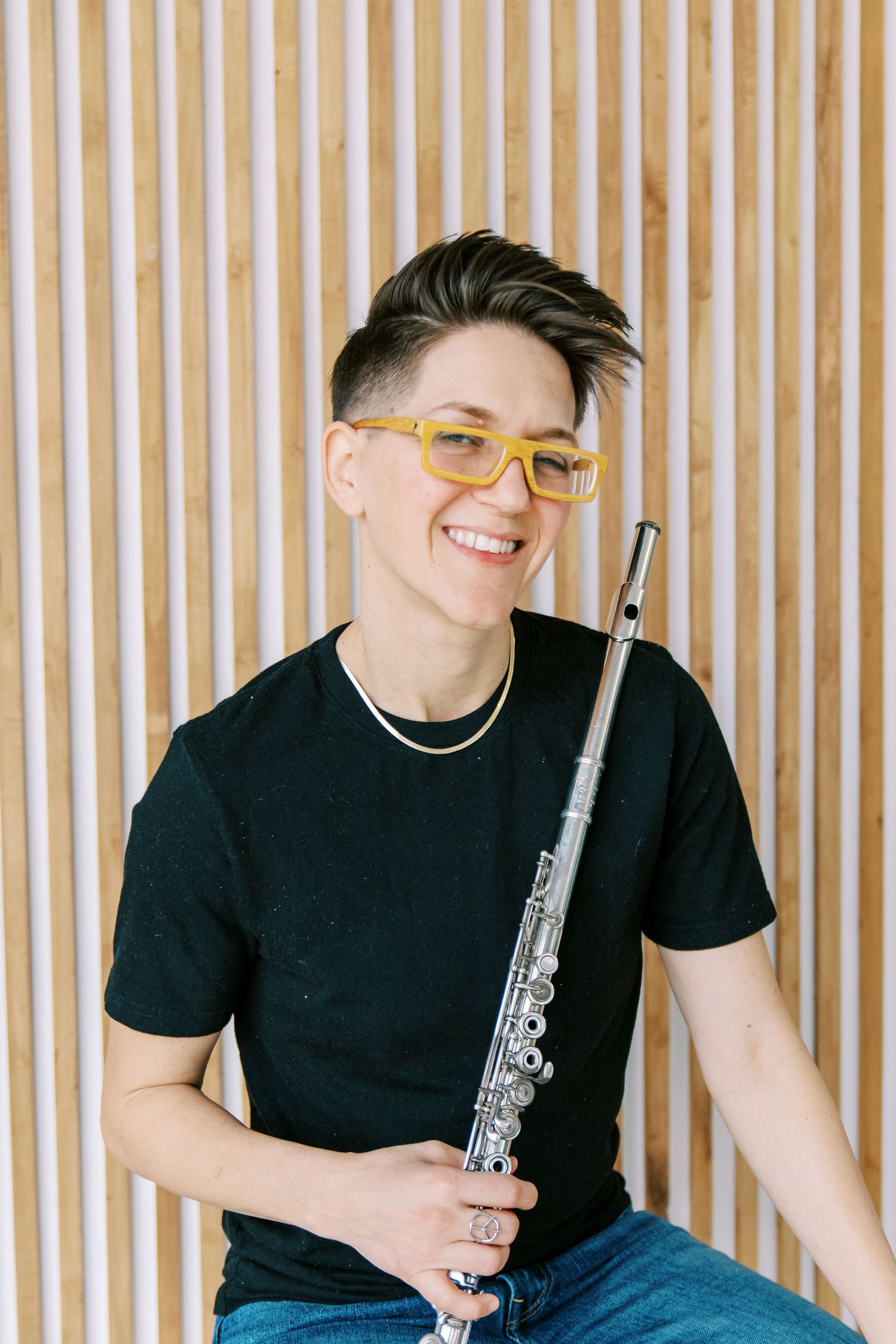 Musician in black t-shirt and jeans holding a flute while sitting against striped wall background.