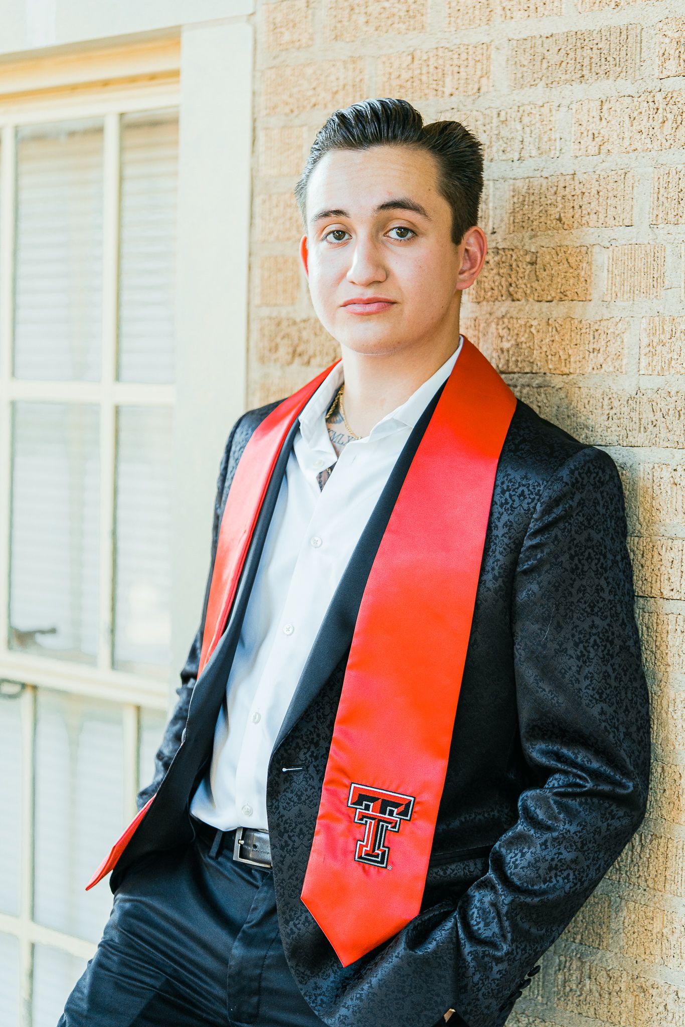 Graduate wearing black suit and red Texas Tech graduation stole stands by brick building.
