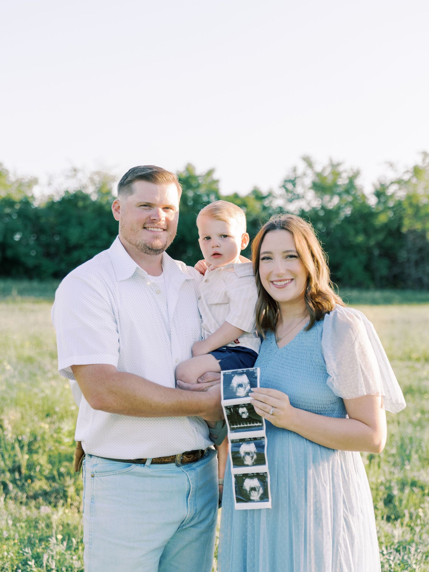 A family in a field holds up ultrasound photos while wearing coordinating blue and white outfits.