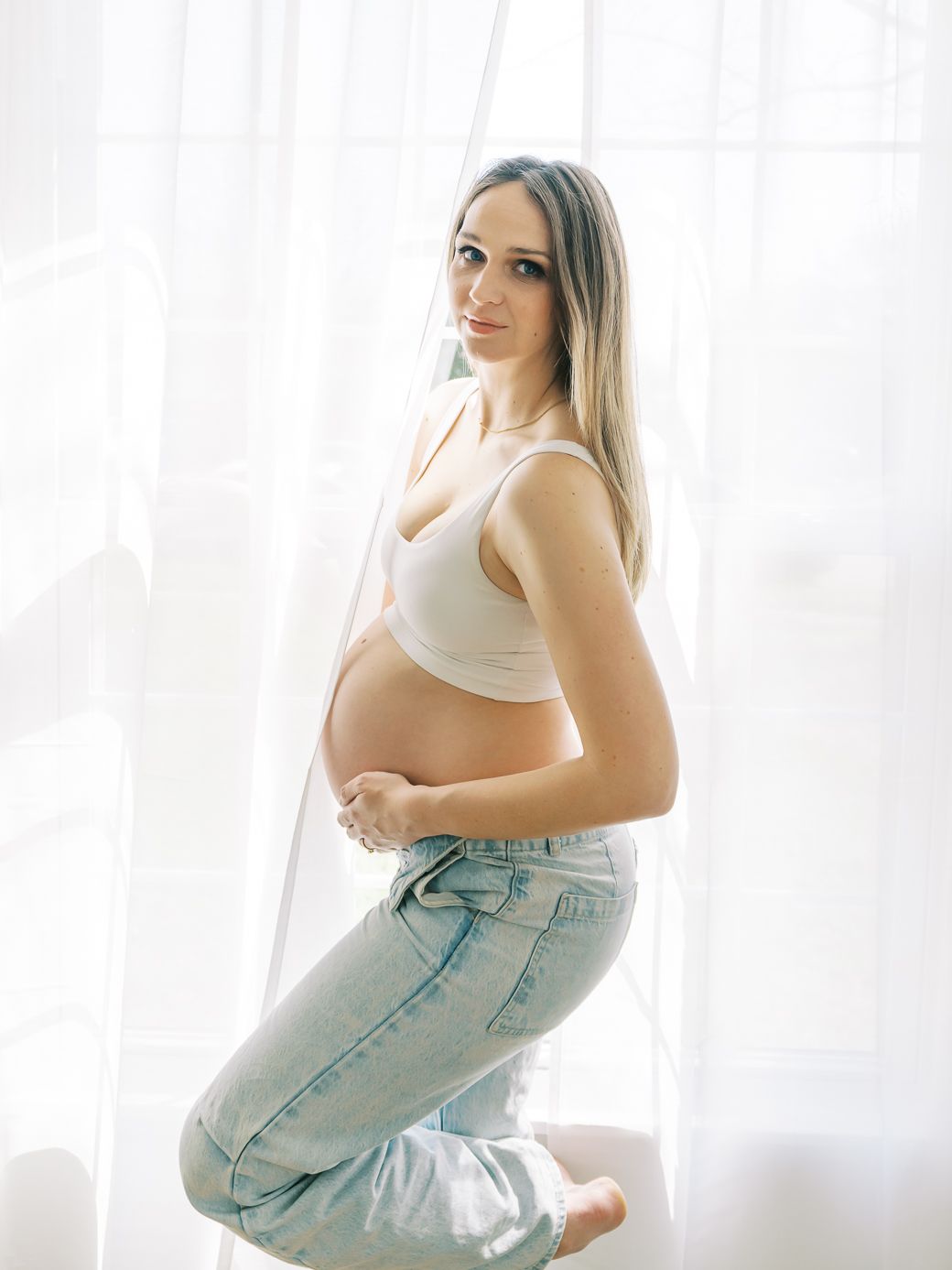 Expectant mother in casual denim and white top poses by window in natural light.