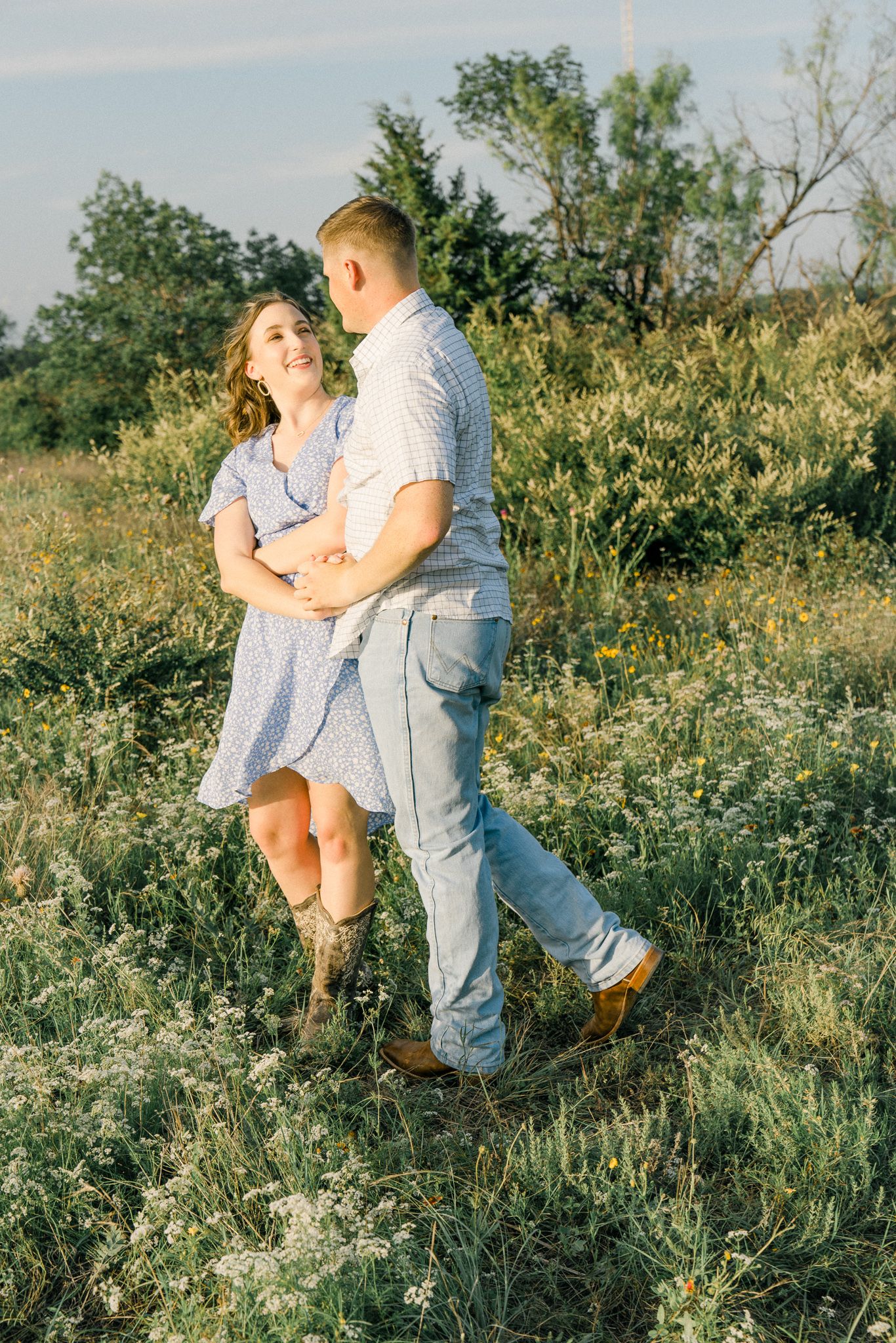 A couple embraces and dances together in a sunlit meadow filled with wildflowers and tall grass.