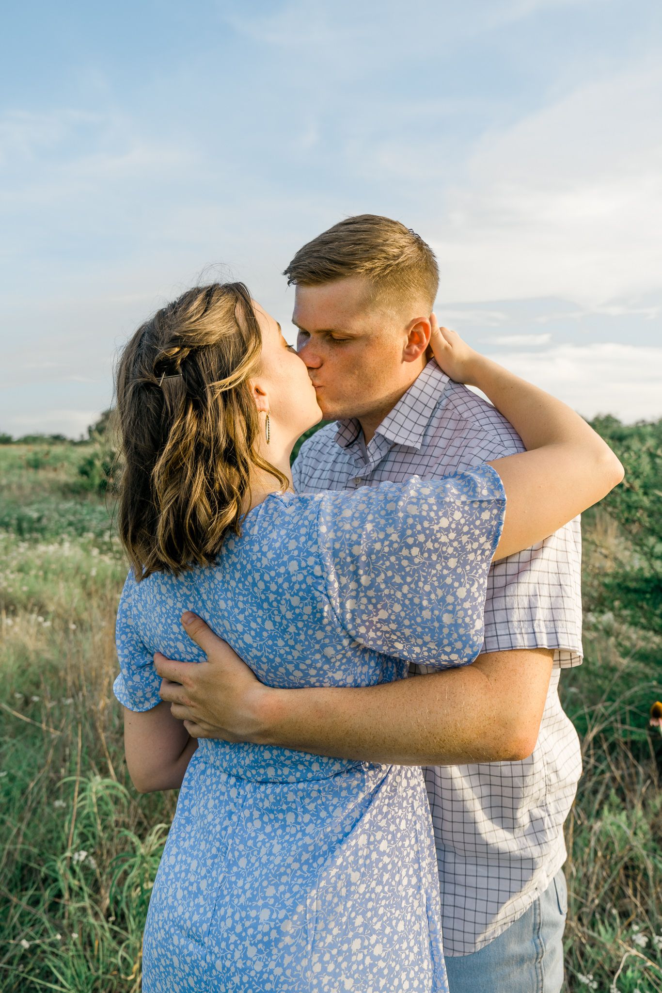 A couple embraces in a sunny field with tall grass and wildflowers during a romantic outdoor photoshoot.