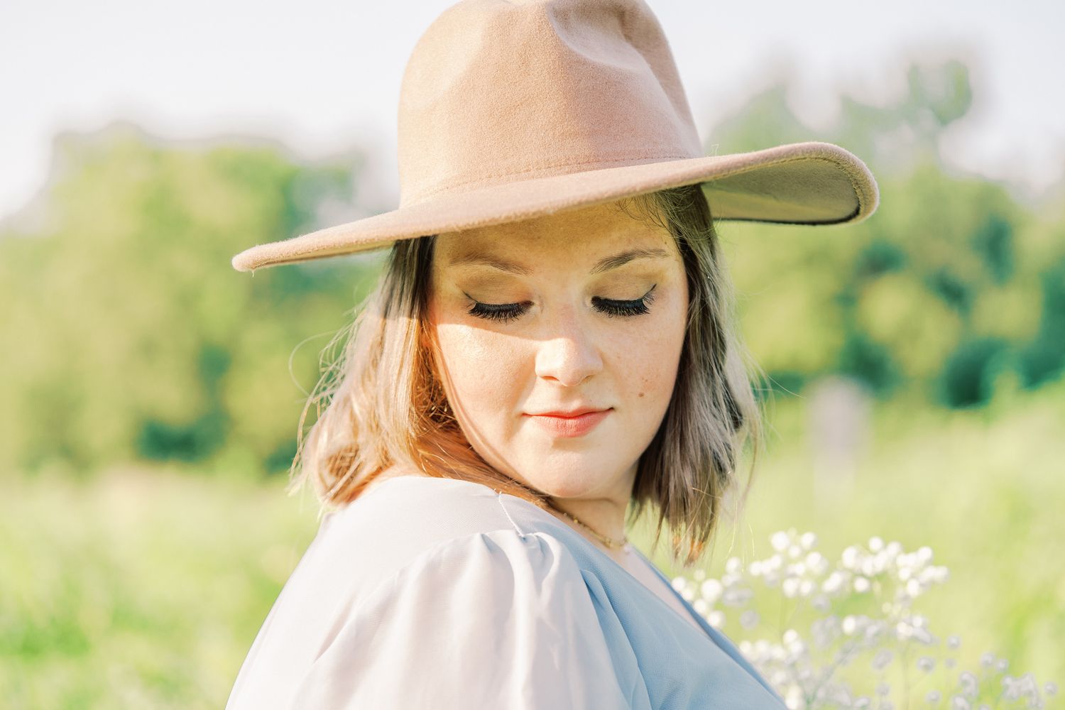 An expectant mother wearing a beige hat and blue dress poses gracefully among summer wildflowers.