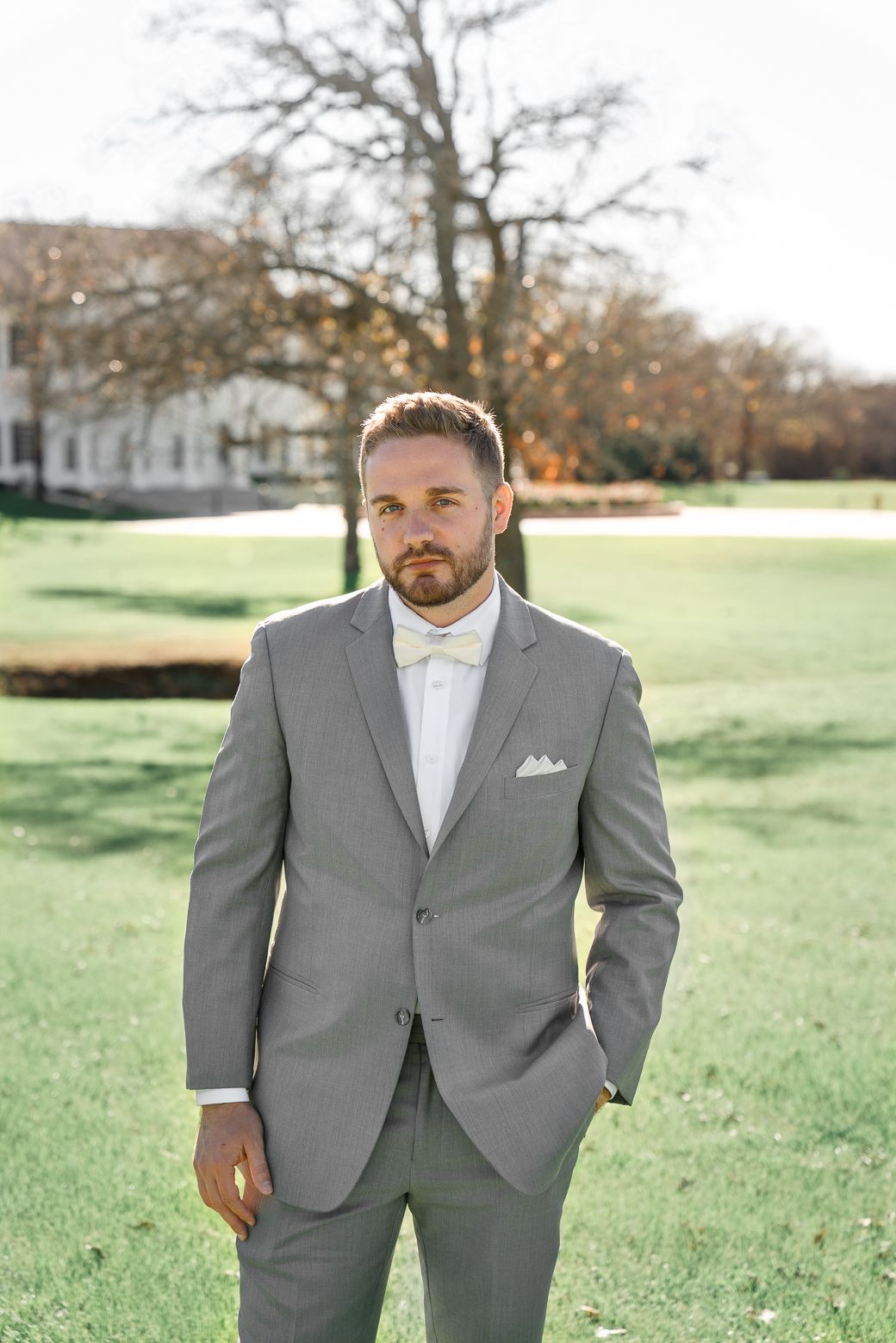 A person in a light gray suit and white shirt poses outdoors on a grassy lawn with autumn trees in the background.
