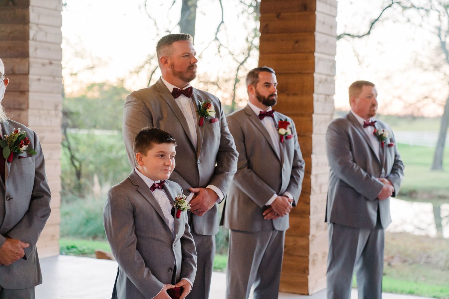 Groomsmen in gray suits and red bowties pose together in a rustic outdoor setting during a wedding celebration.