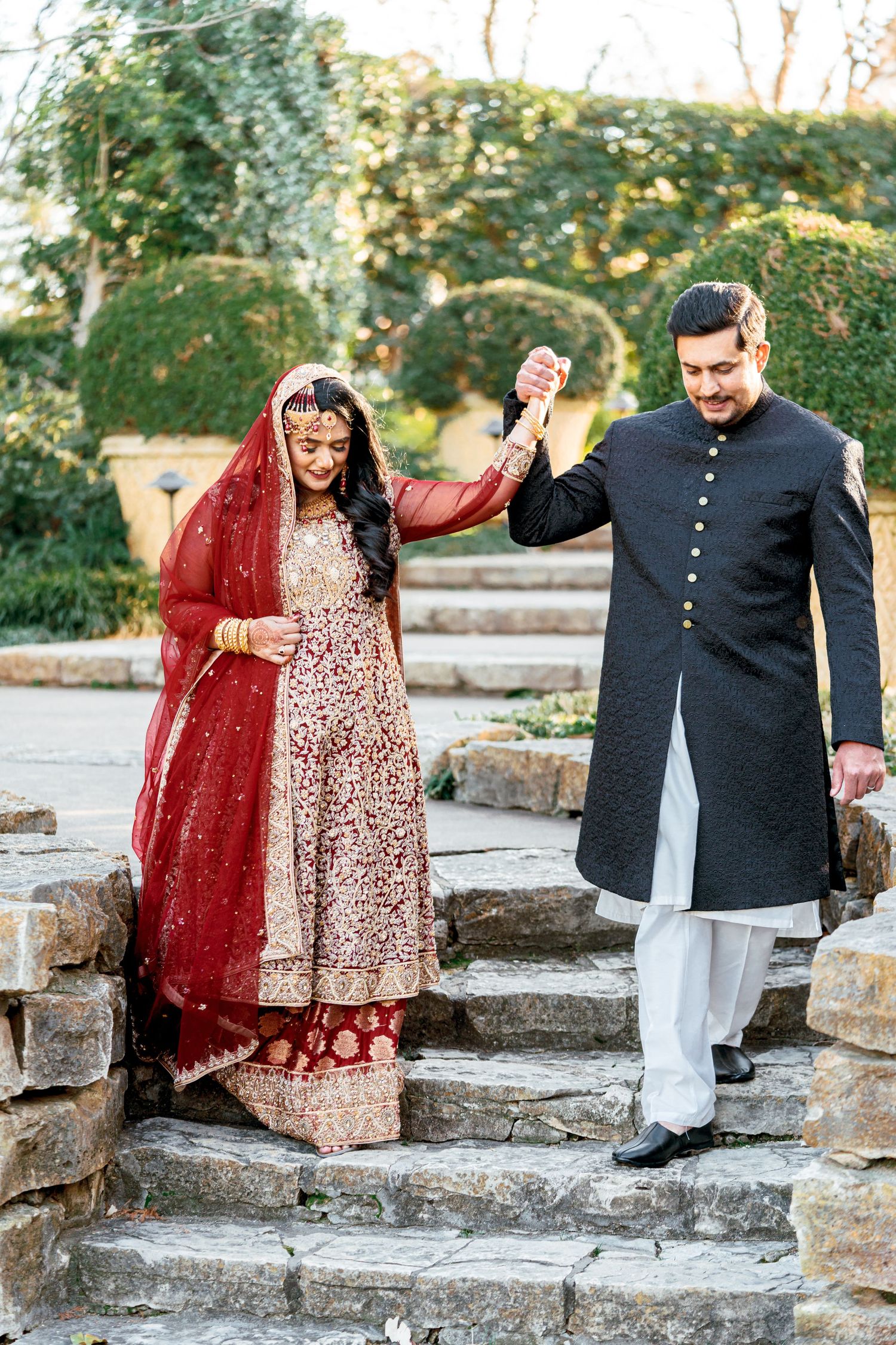 A couple in traditional South Asian wedding attire playfully posing on stone steps outdoors.
