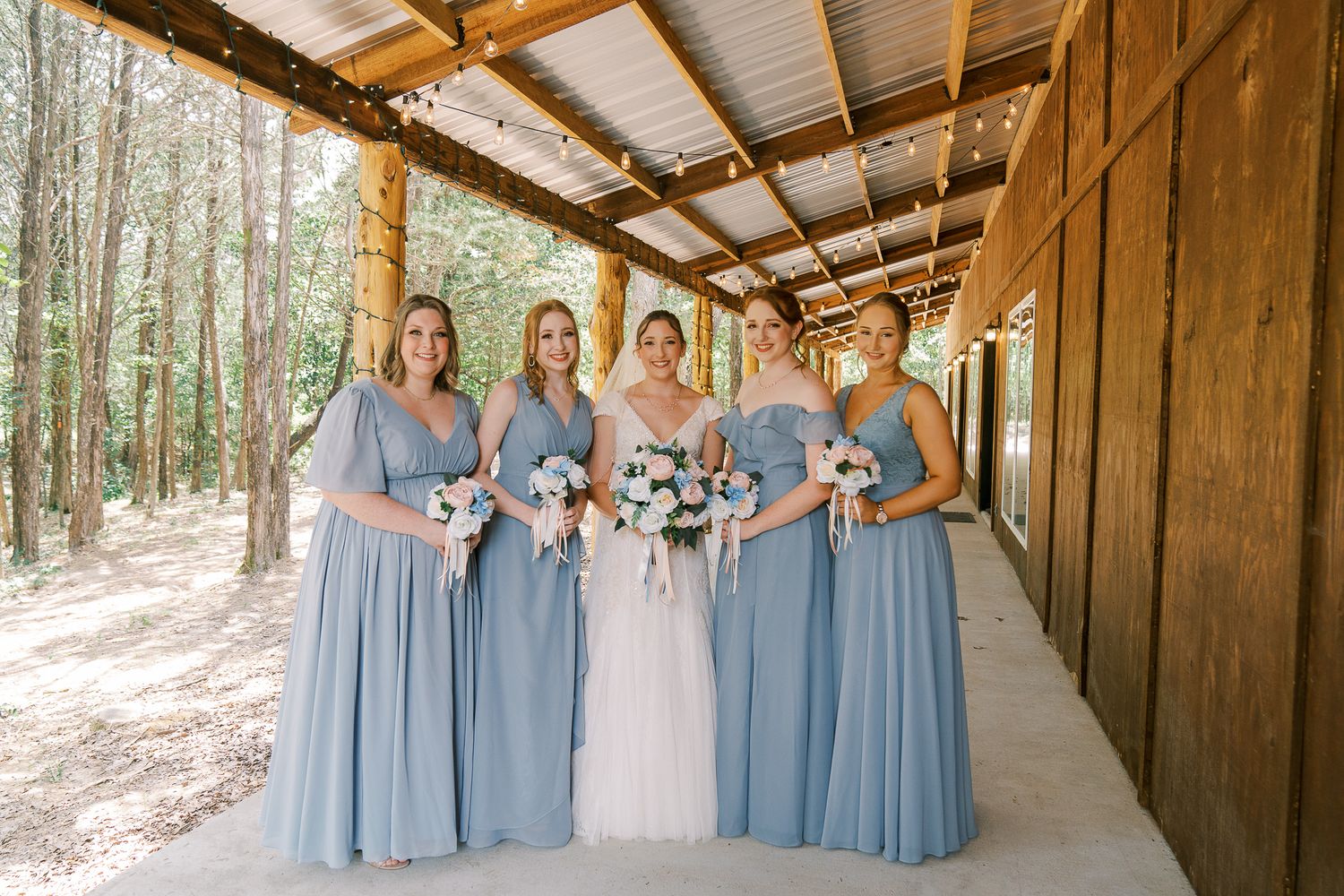 A bridal party poses together in a rustic wooden covered walkway surrounded by trees.