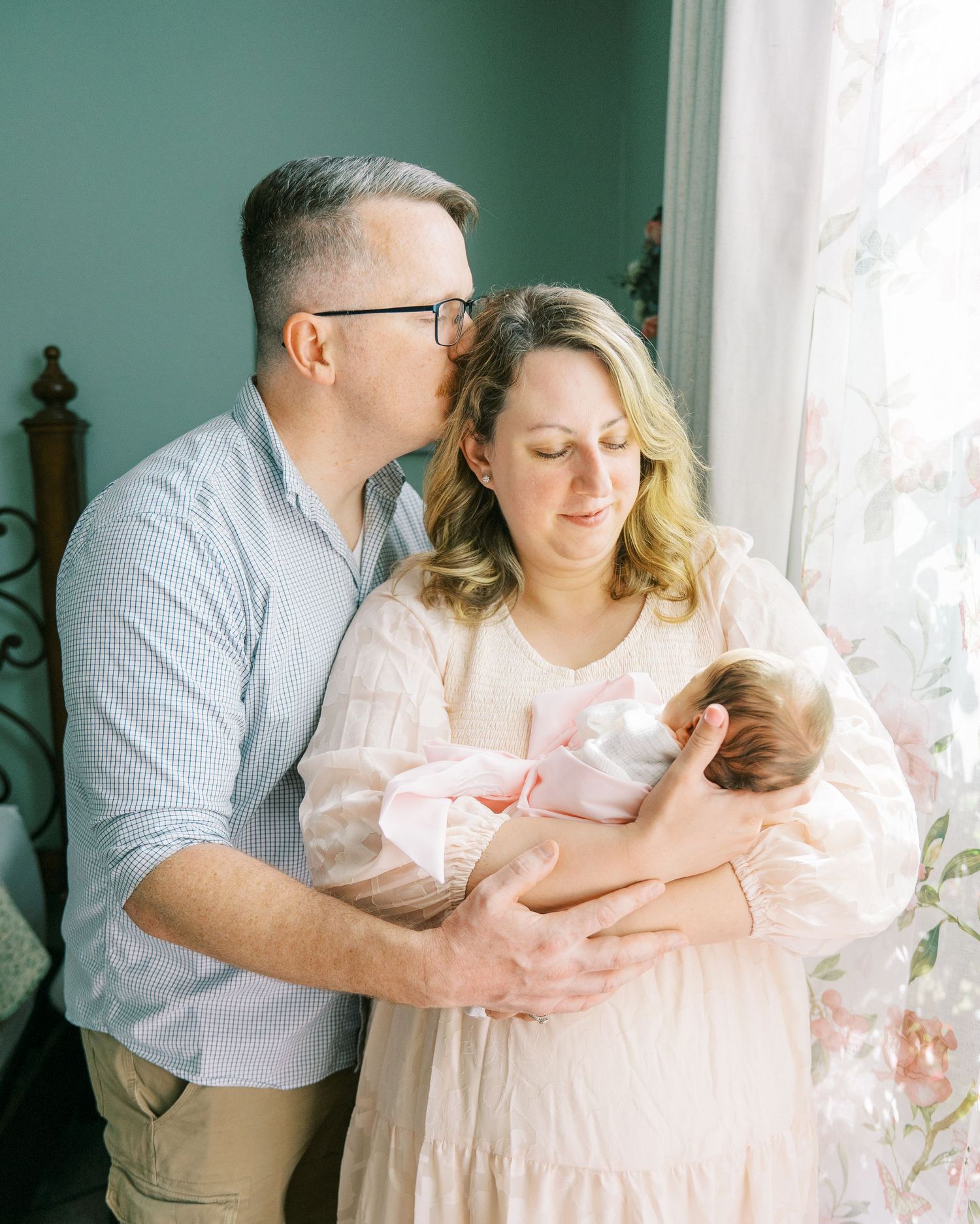 A tender family moment as parents cradle their newborn baby near a sunlit window with sheer curtains.