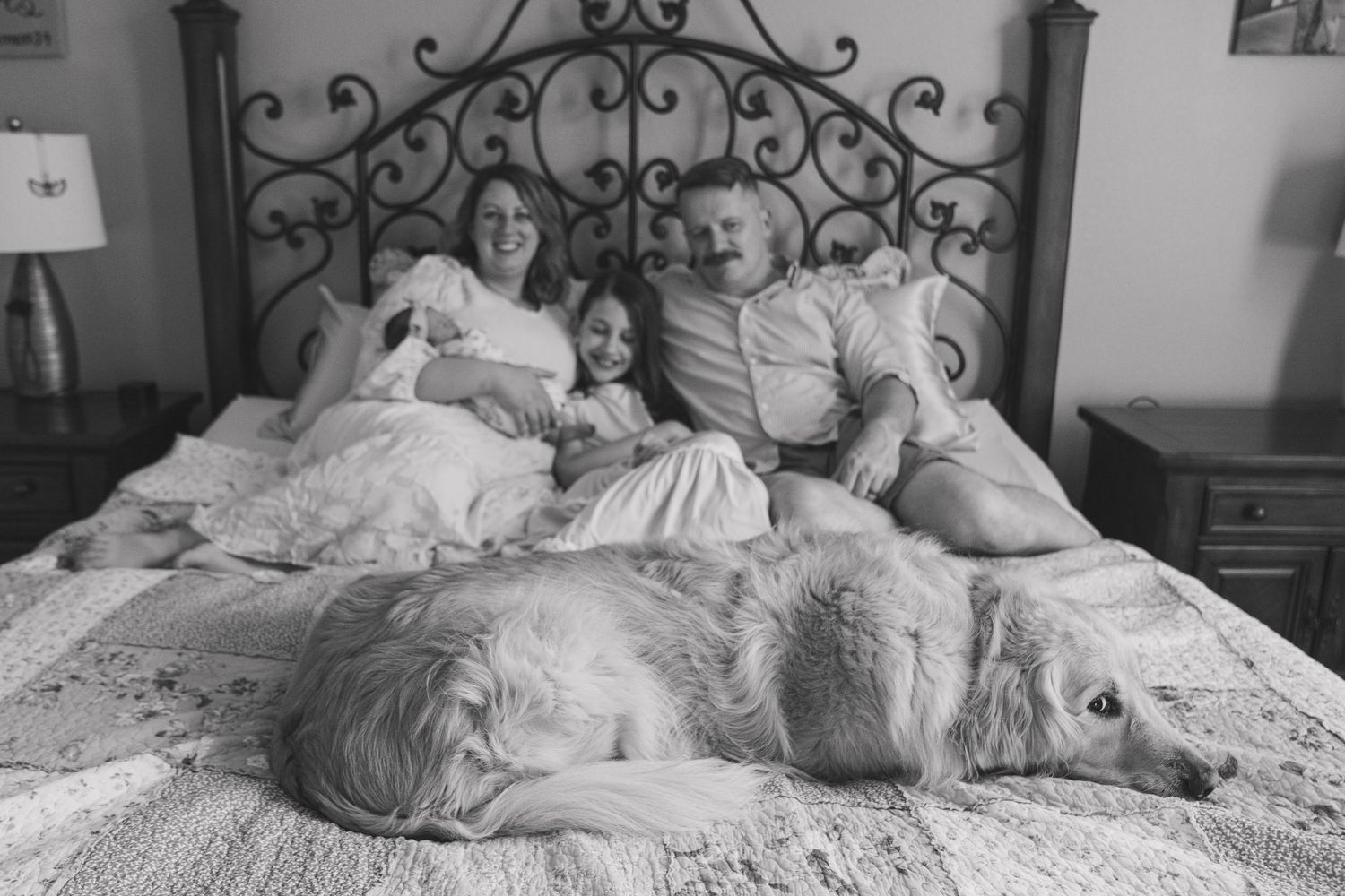 Black and white photo of family and pets lounging together on a bed with ornate headboard.