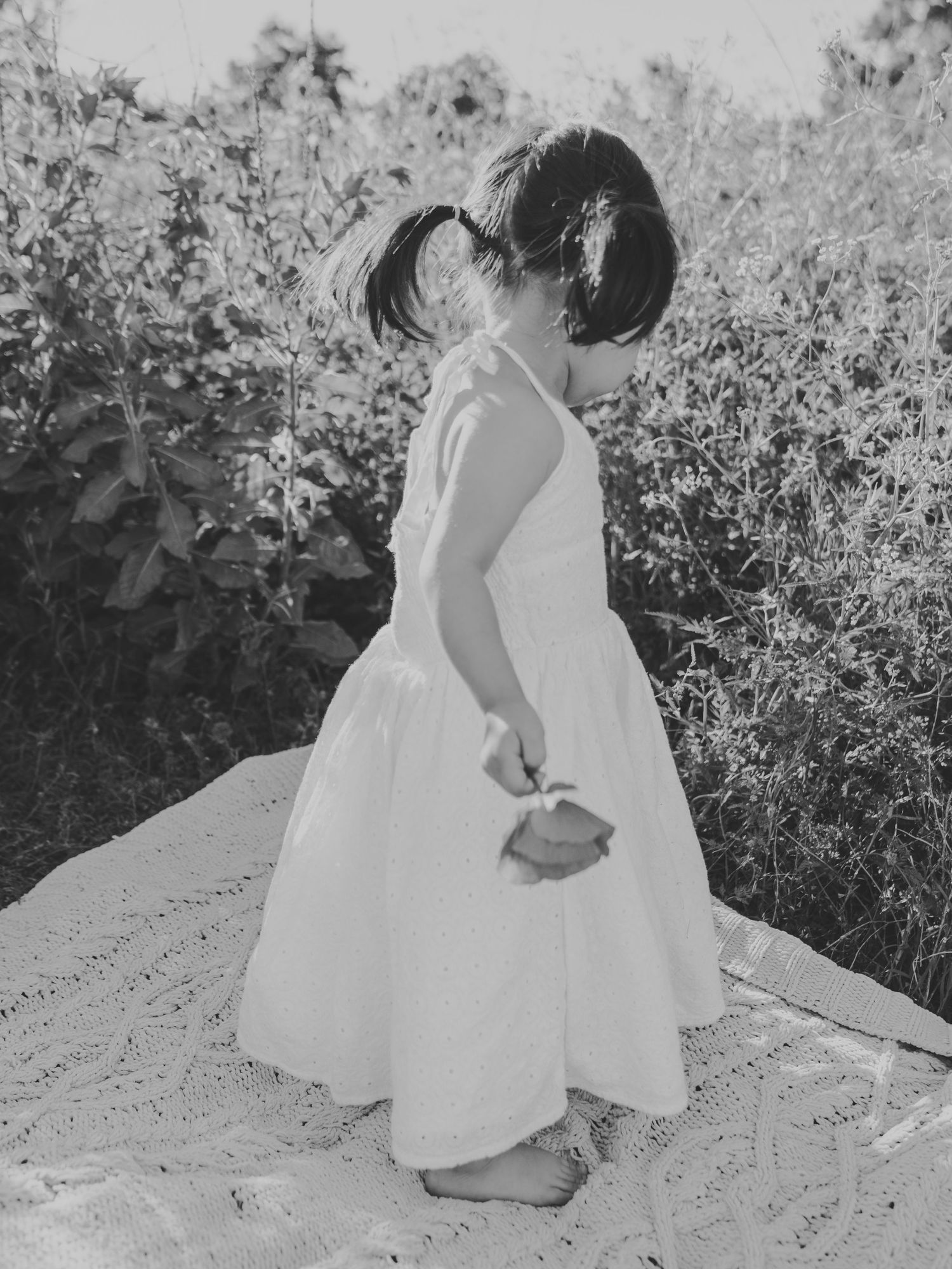 Black and white photograph of a small child in a white dress twirling outdoors among flowering bushes.