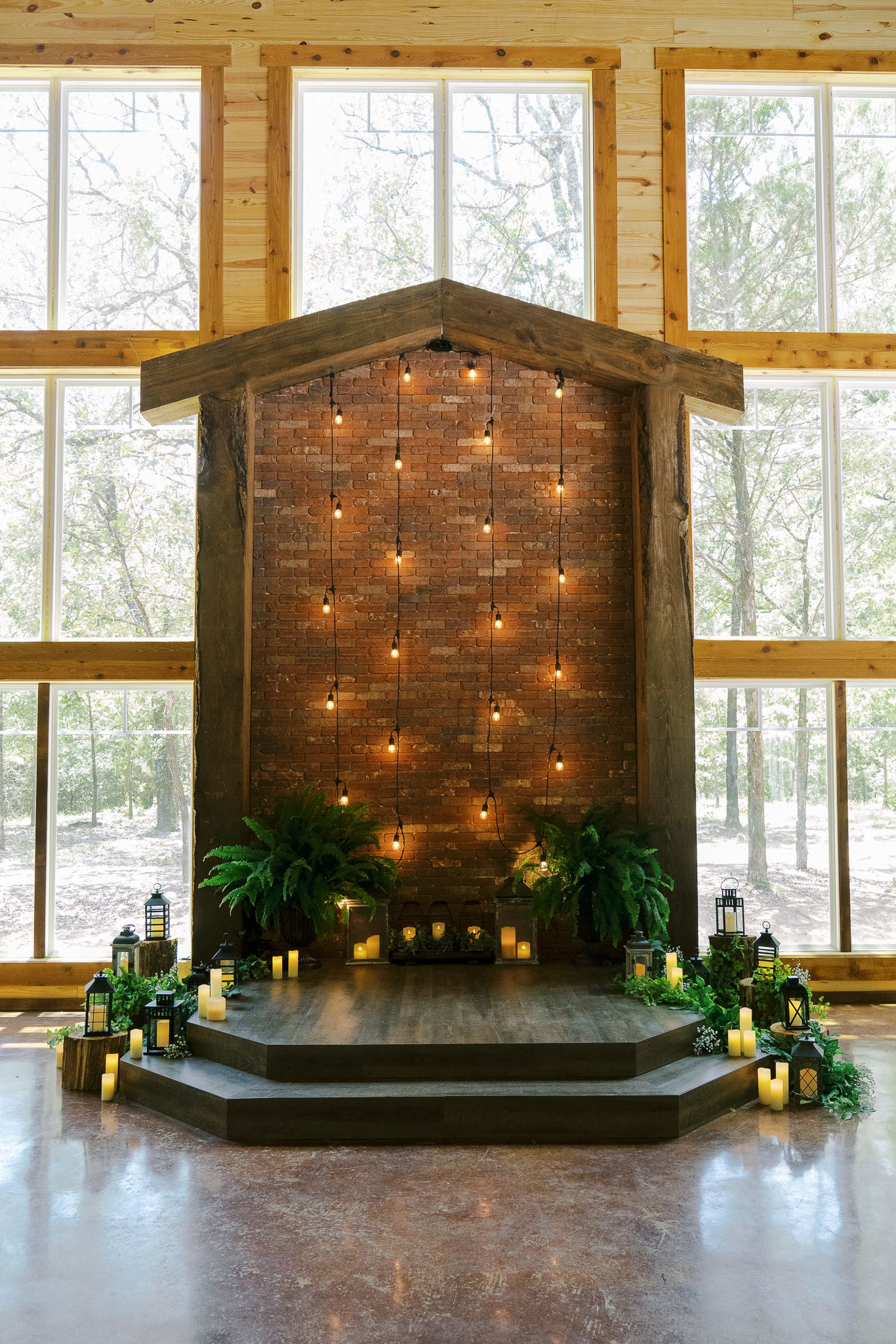 Rustic wooden event venue stage decorated with string lights, lanterns and greenery against large windows overlooking nature.