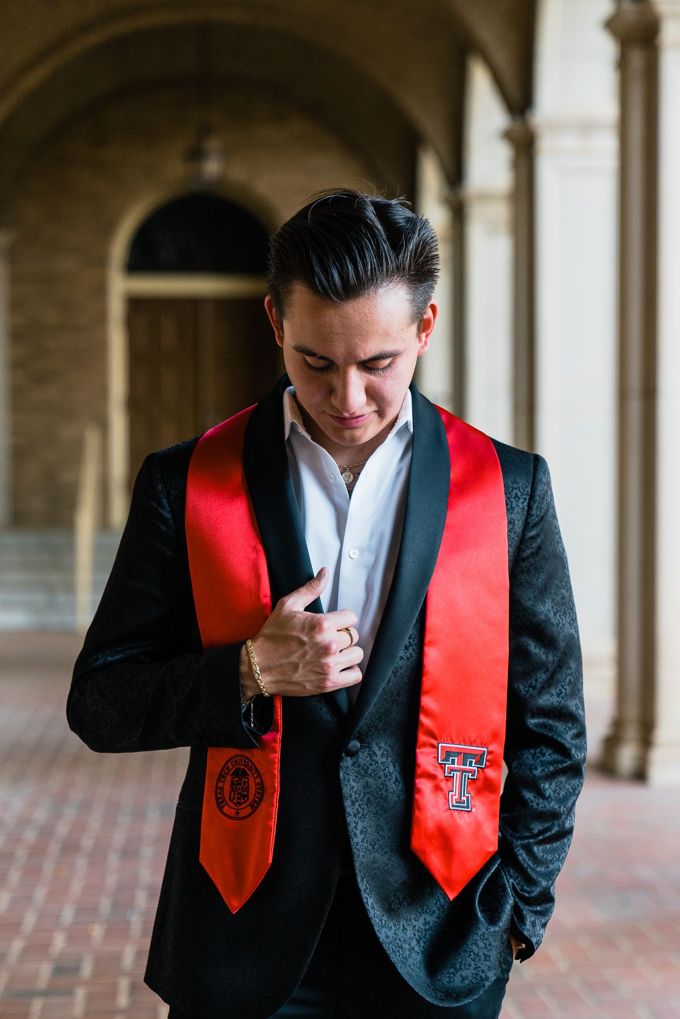 Graduate wearing black suit and red graduation stoles stands thoughtfully in arched hallway.