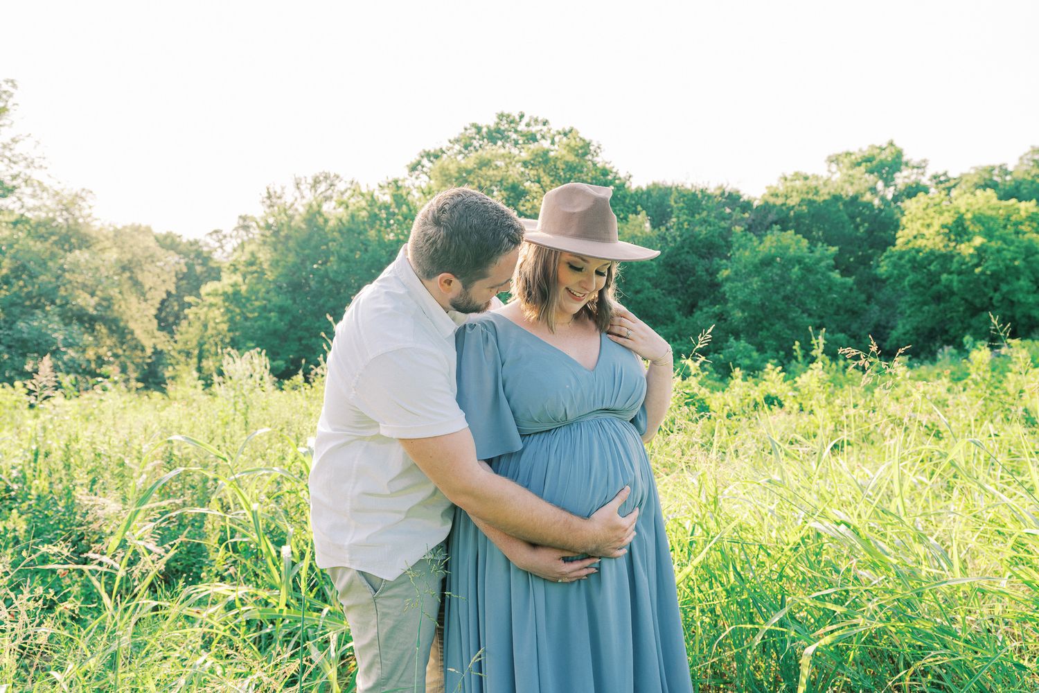 A couple embraces in a sunlit meadow surrounded by tall grass and wildflowers during golden hour.
