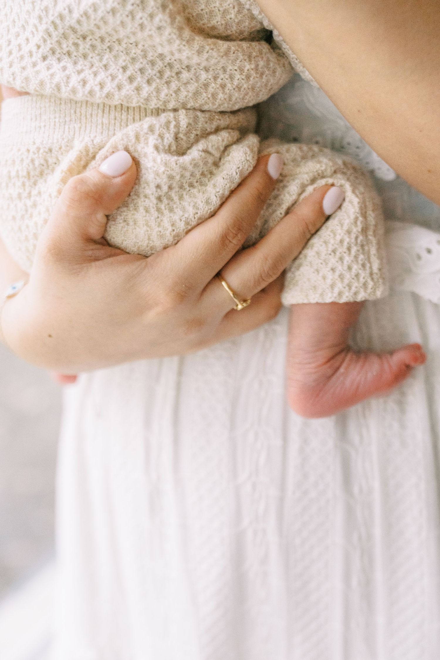 A mother gently holds her newborn baby wrapped in a beige knit blanket, wearing a delicate gold ring.