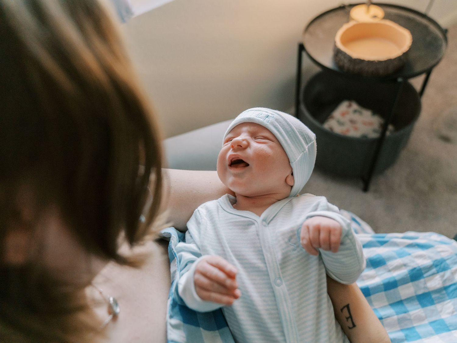 Newborn baby in blue outfit and hat crying while being held by mother in warm nursery setting.