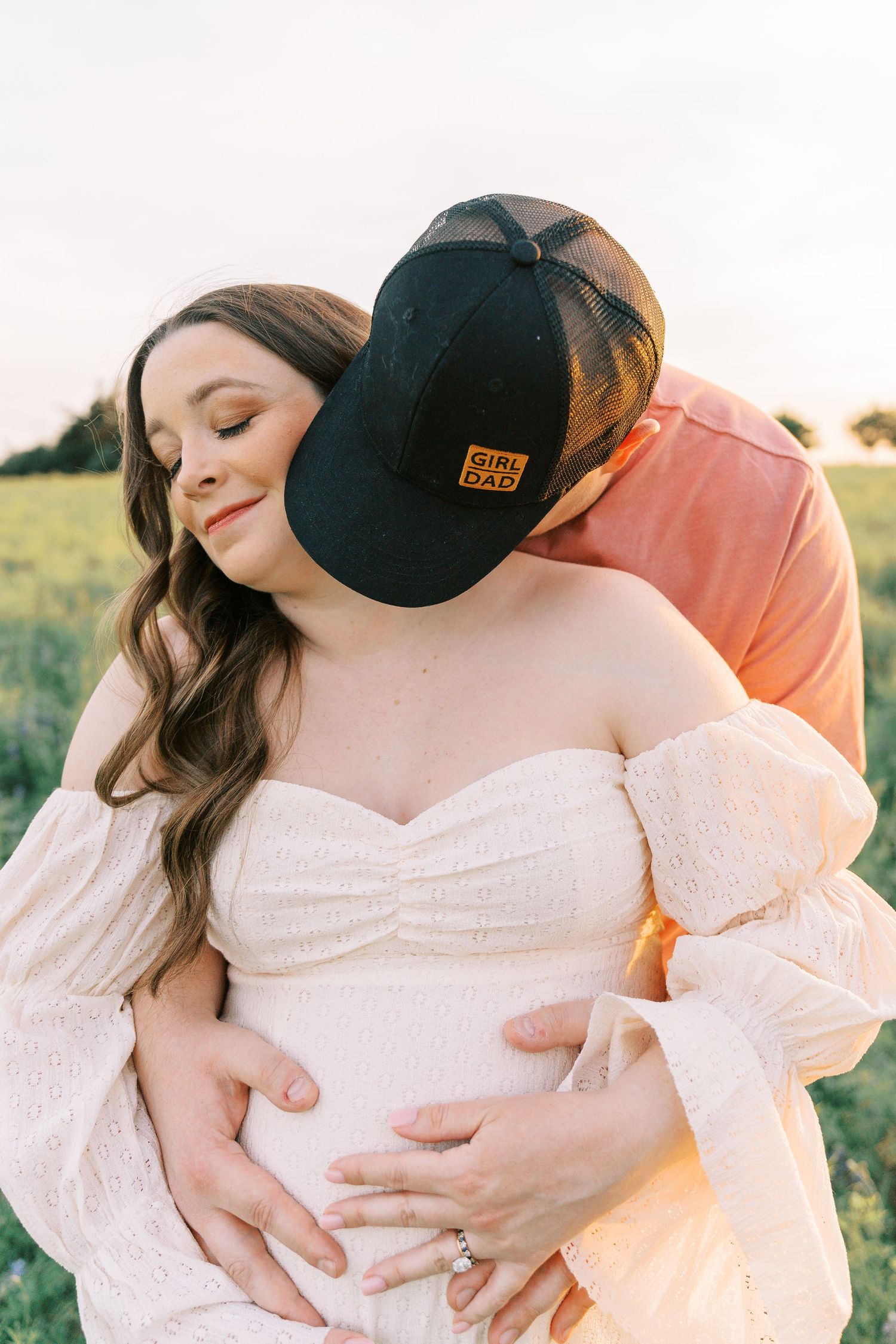 Couple sharing a tender maternity moment outdoors, man in Girl Dad cap embracing pregnant woman in white dress at golden hour.