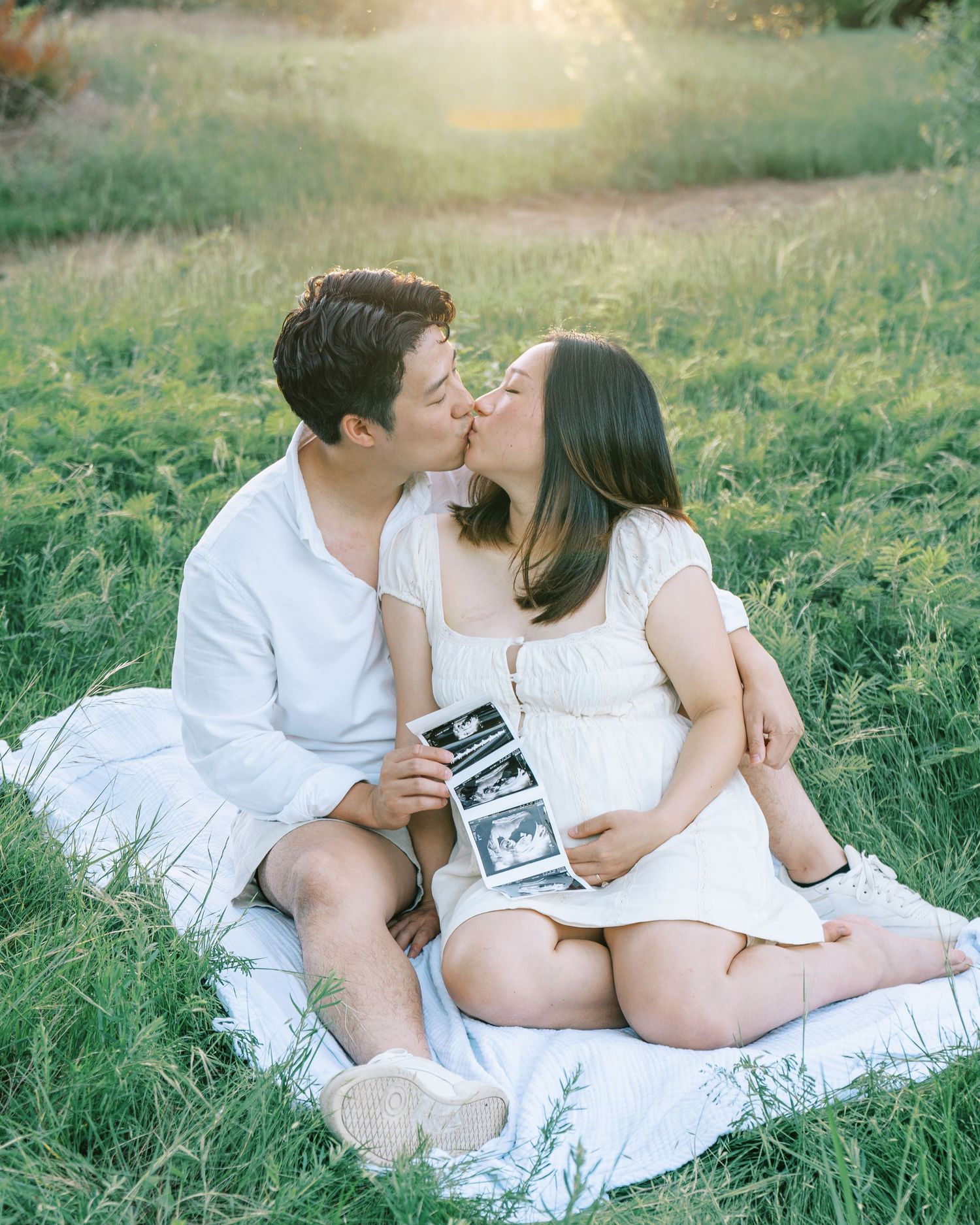 Couple kissing on white blanket in sunlit field, holding ultrasound photos, maternity announcement portrait.