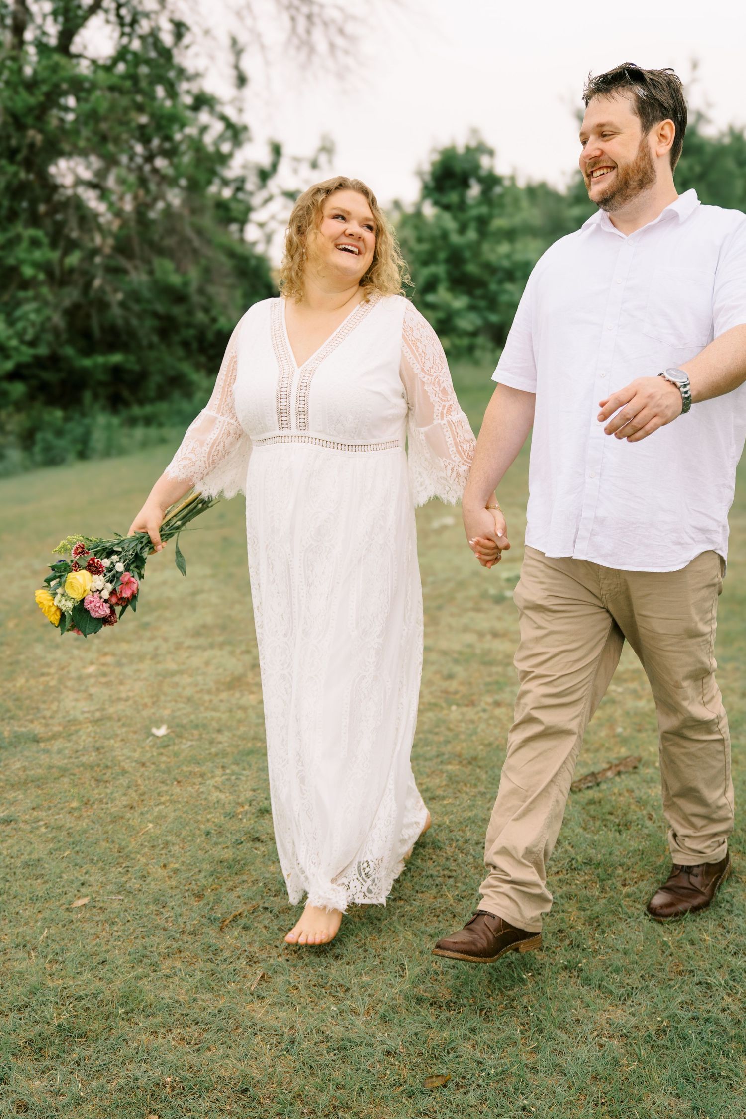 Laughing couple walking hand in hand outdoors, woman in white lace dress holding colorful bouquet.