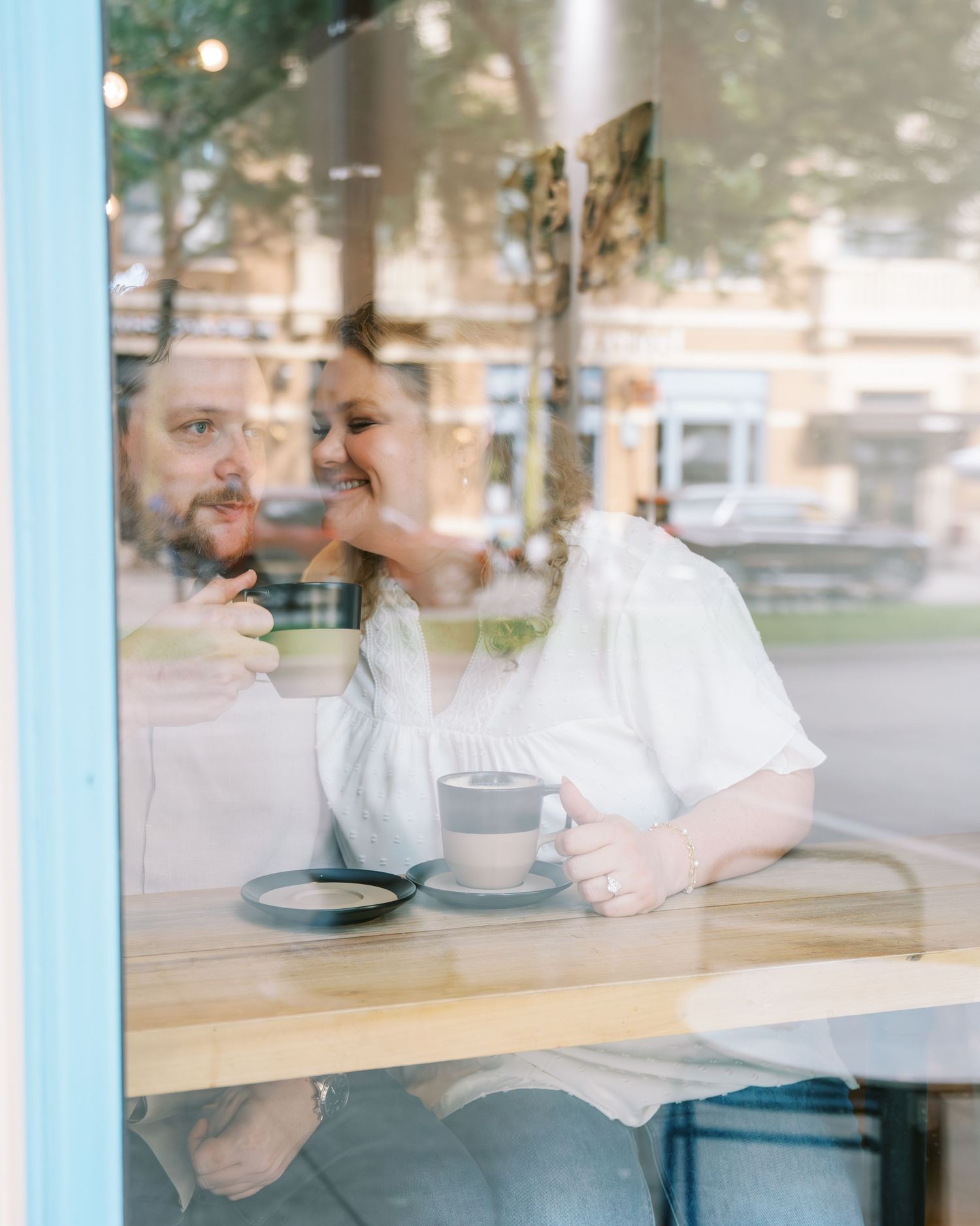 Couple photographed through a cafe window, smiling and holding coffee cups at a wooden table.