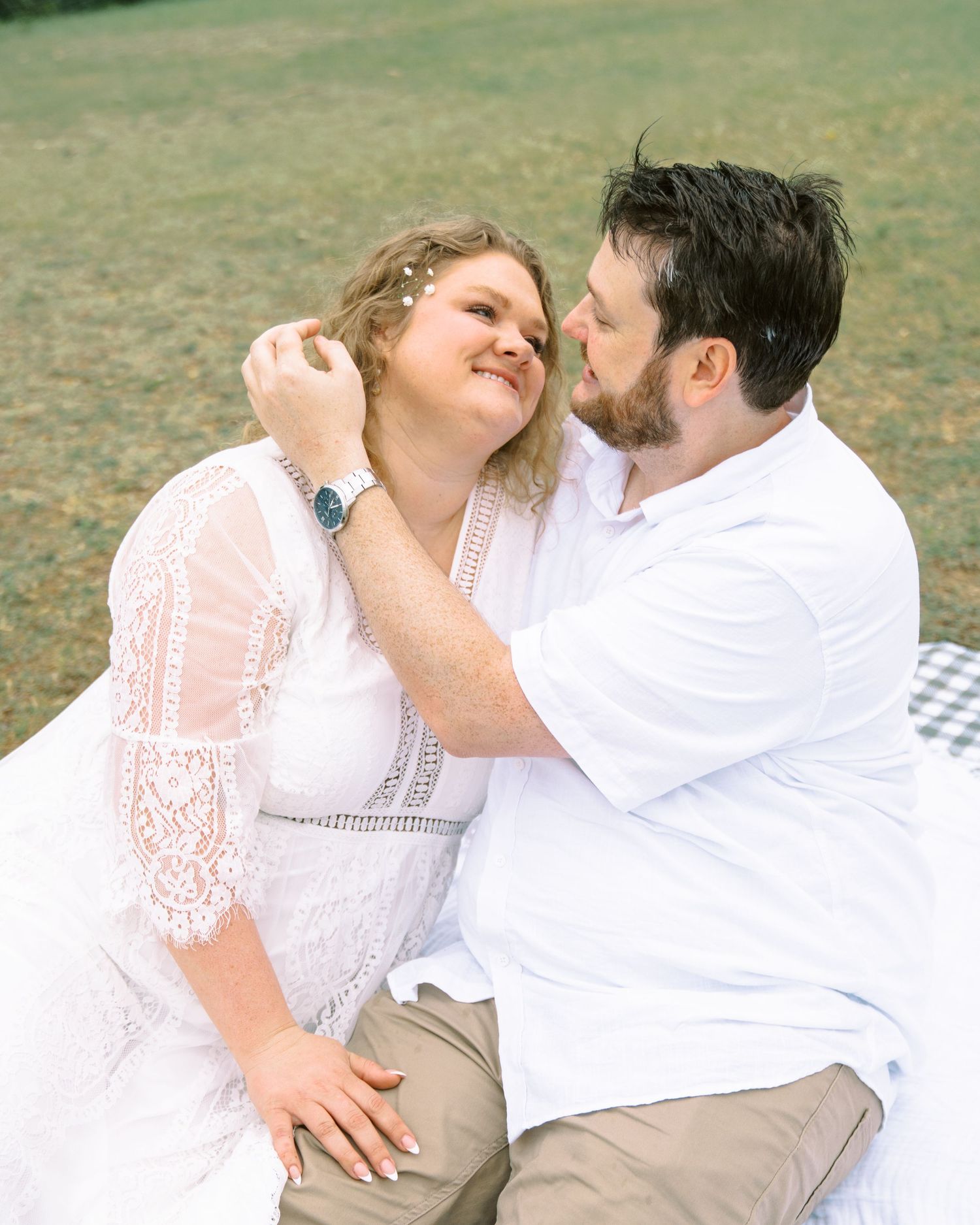 Couple sitting on blanket outdoors, man tenderly touching womans face as they smile at each other.