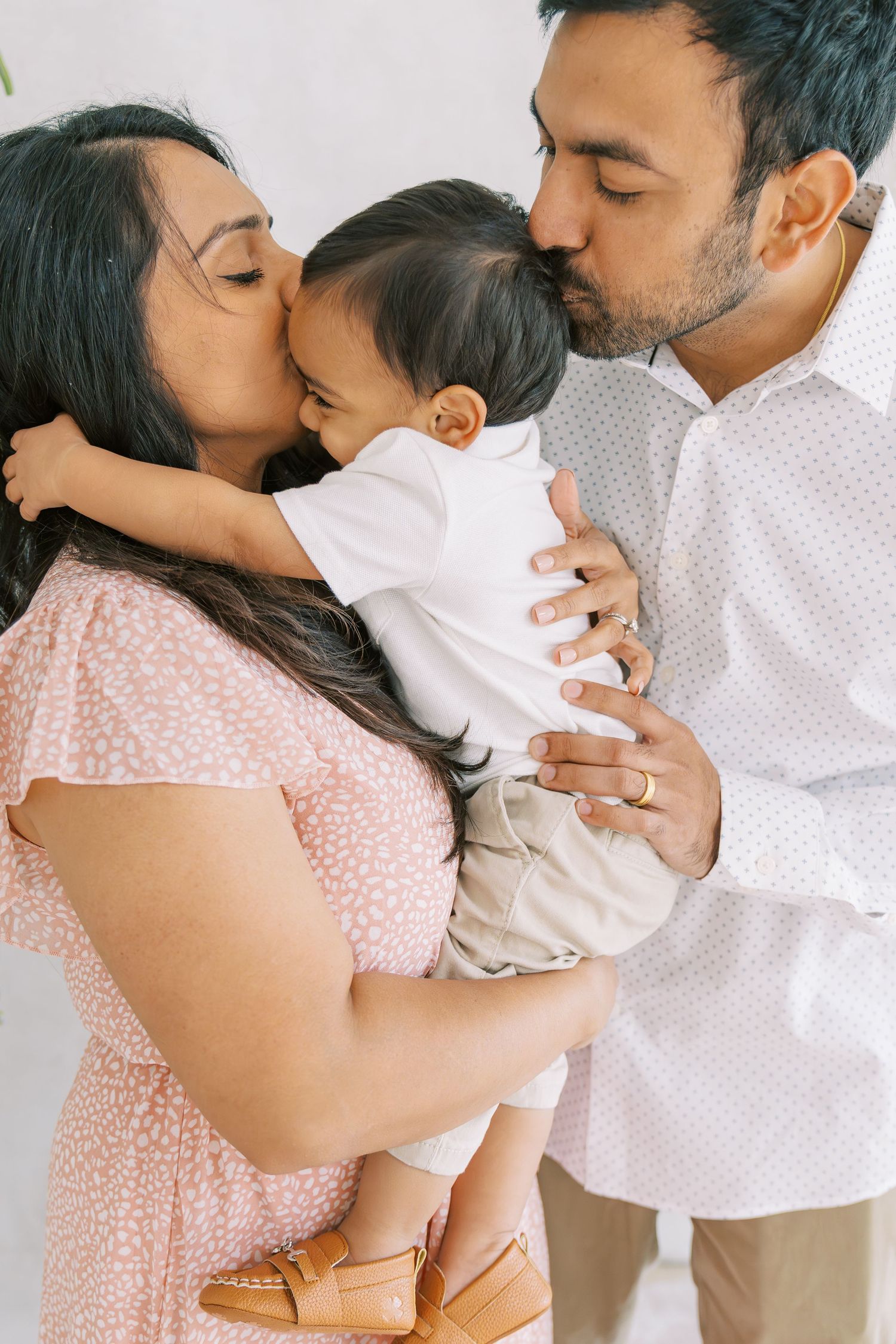 Parents kissing their baby on the cheeks, mother in pink dress, father in white shirt, family portrait.