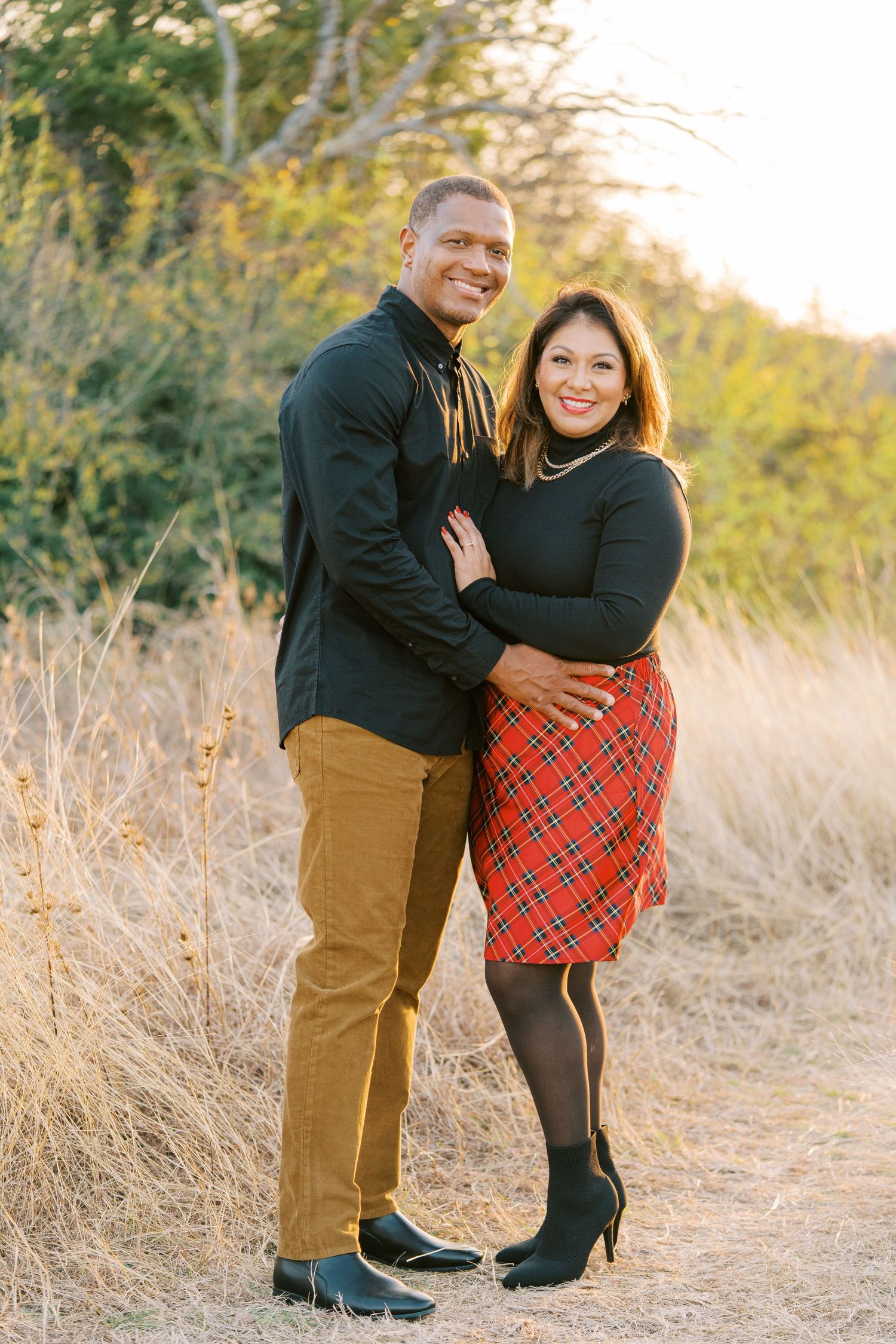 Couple posing outdoors in autumn field, man in black shirt and tan pants, woman in red plaid skirt and black turtleneck.