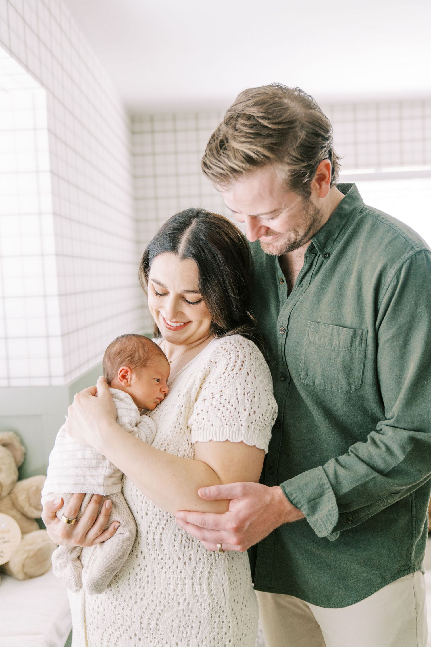 A couple in white and green clothing share tender moments with their newborn baby in a bright, minimalist setting.
