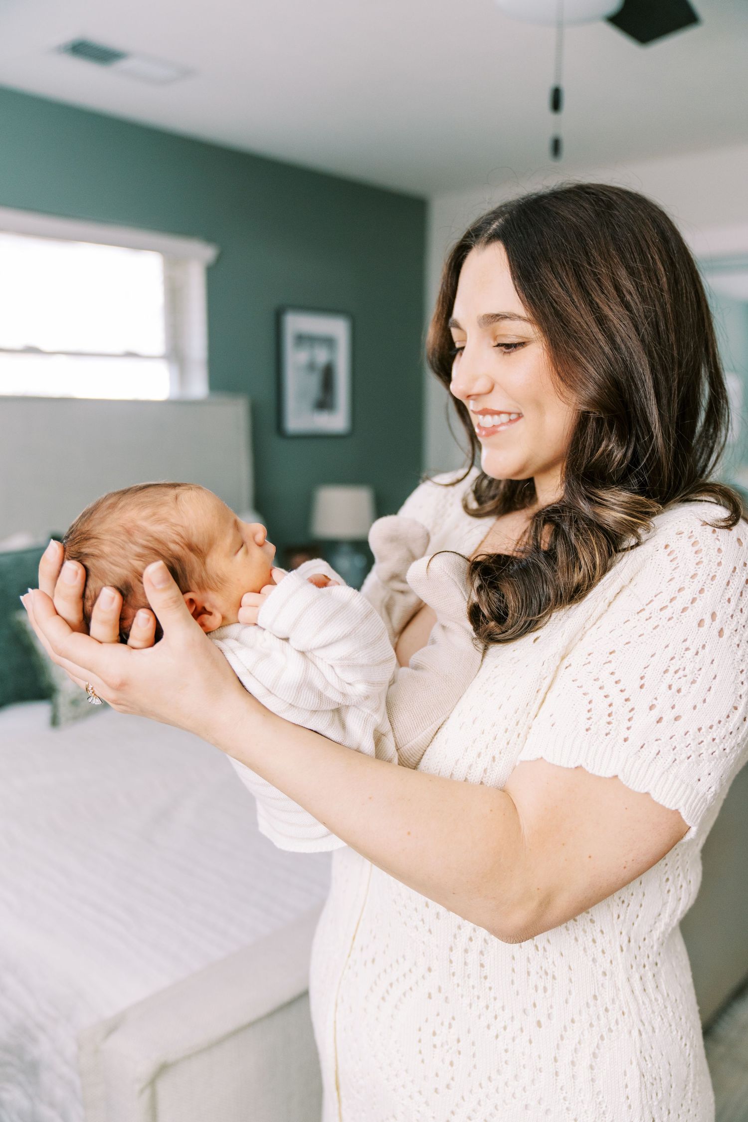 A tender moment captured in a bright bedroom as a mother holds her newborn baby against white bedding.