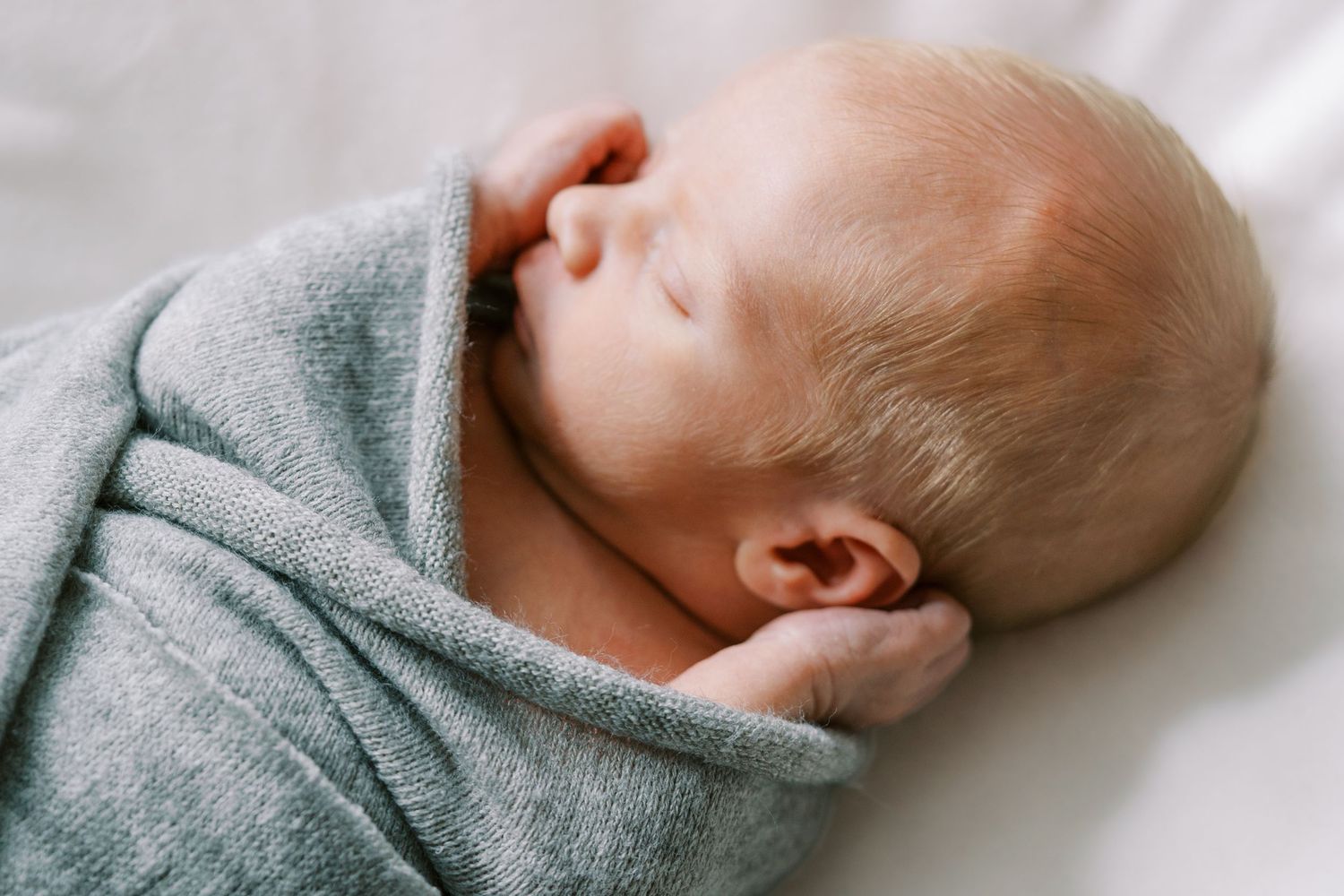 A newborn baby sleeping peacefully on white bedding while wearing a soft gray sweater.