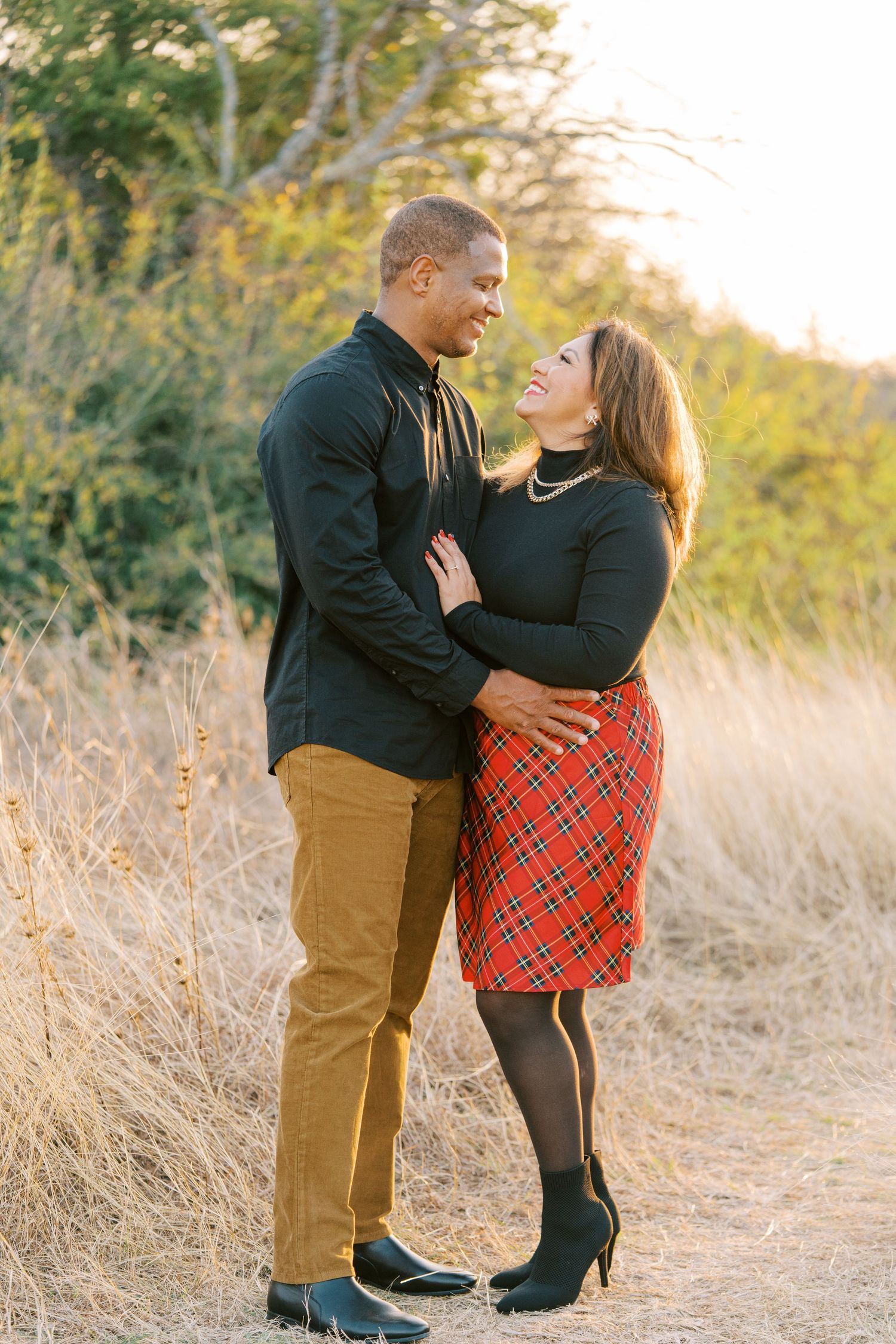 A couple poses together outdoors in a field during golden hour wearing coordinated casual outfits.