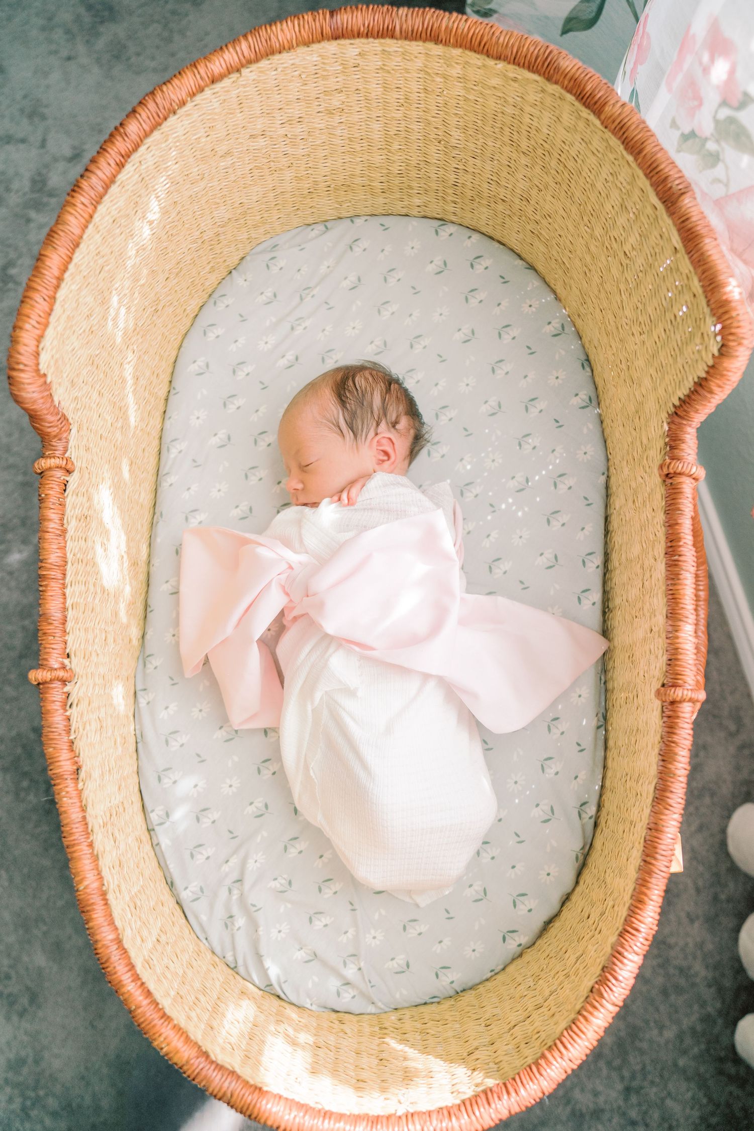 Sleeping newborn in pink outfit resting in round woven basket with gray cushioning.