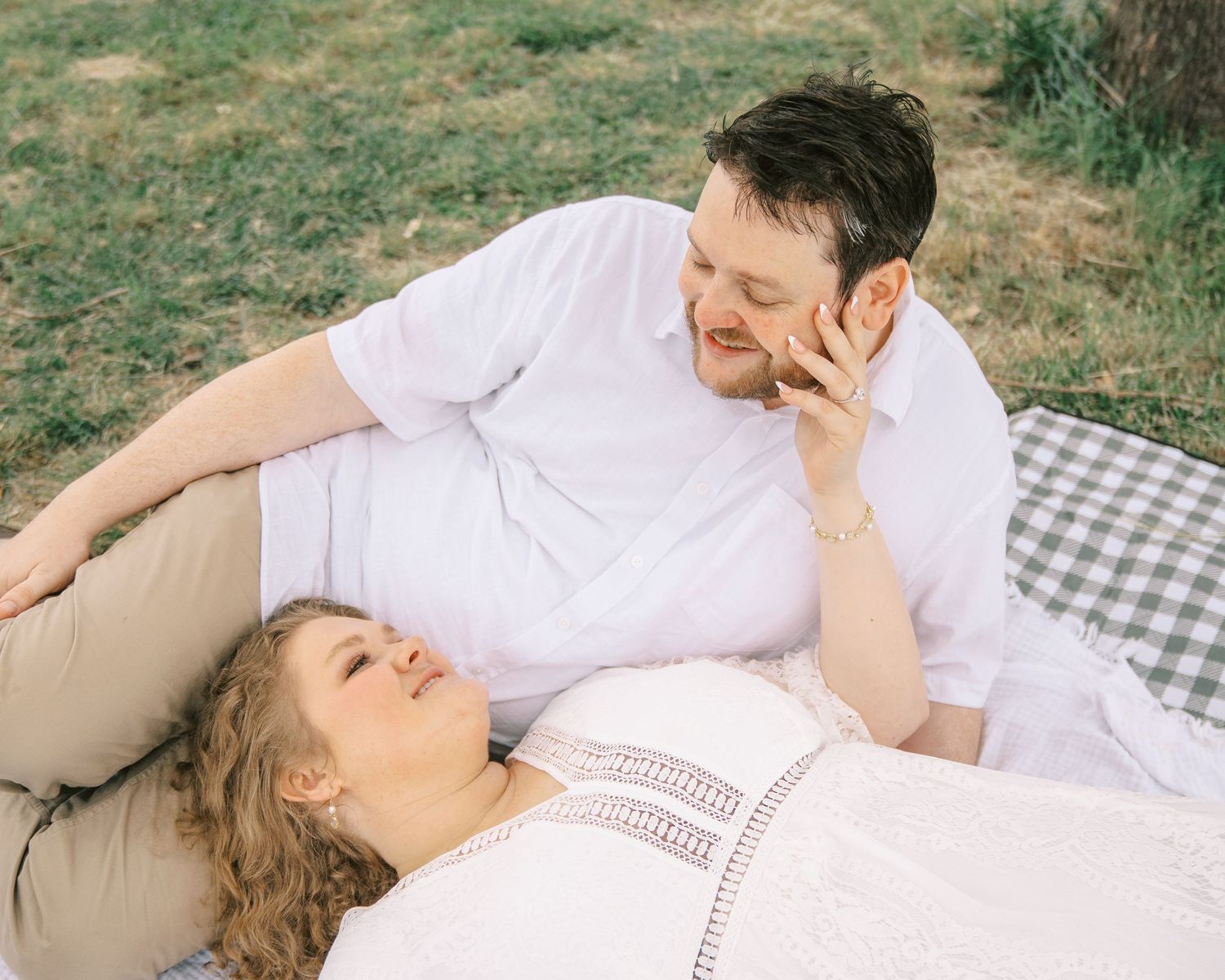 Couple relaxing on a picnic blanket outdoors, woman lying down smiling up at man in white shirt.