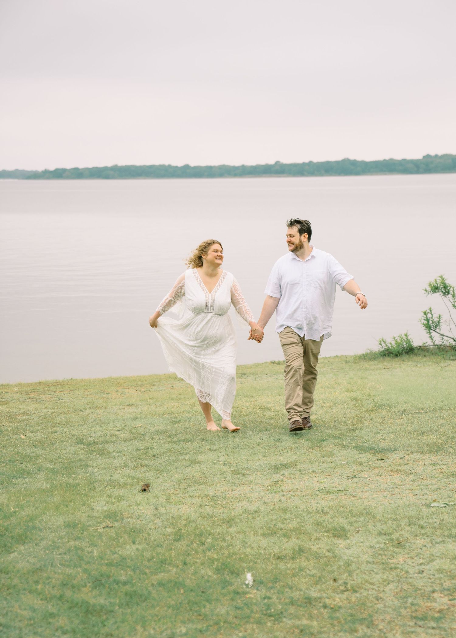 A joyful couple holds hands while walking on green grass by a serene lake, woman in white dress, man in casual attire.