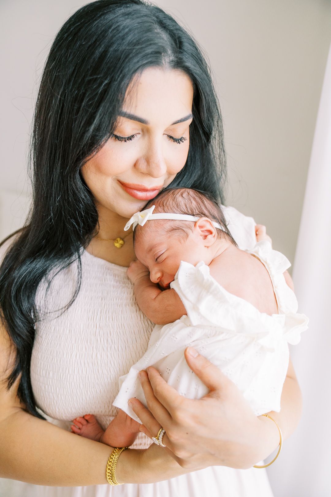 A woman with dark hair in a white dress tenderly holds her sleeping newborn baby wrapped in white cloth.