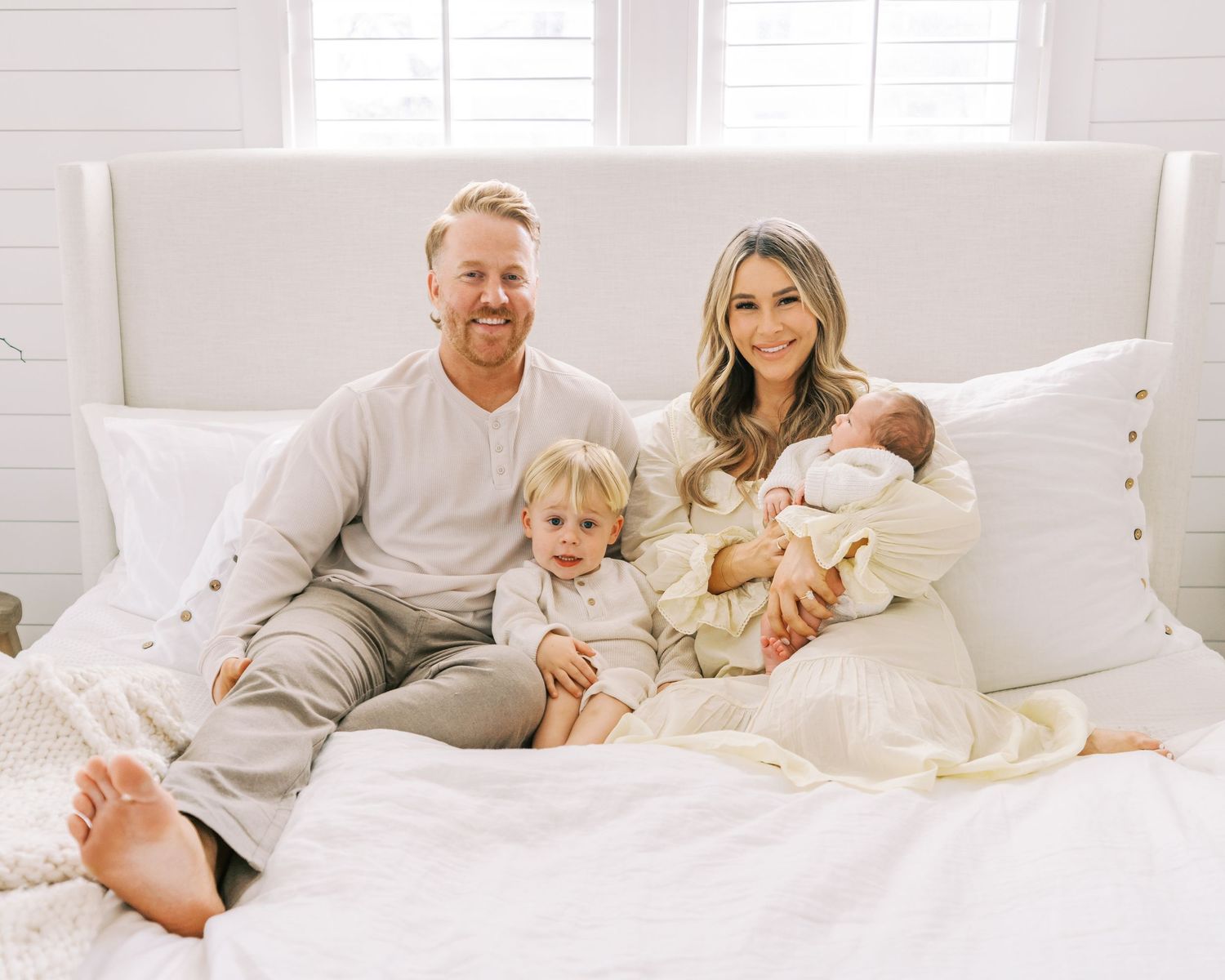 A family of four dressed in neutral colors cuddles together on a white bed.