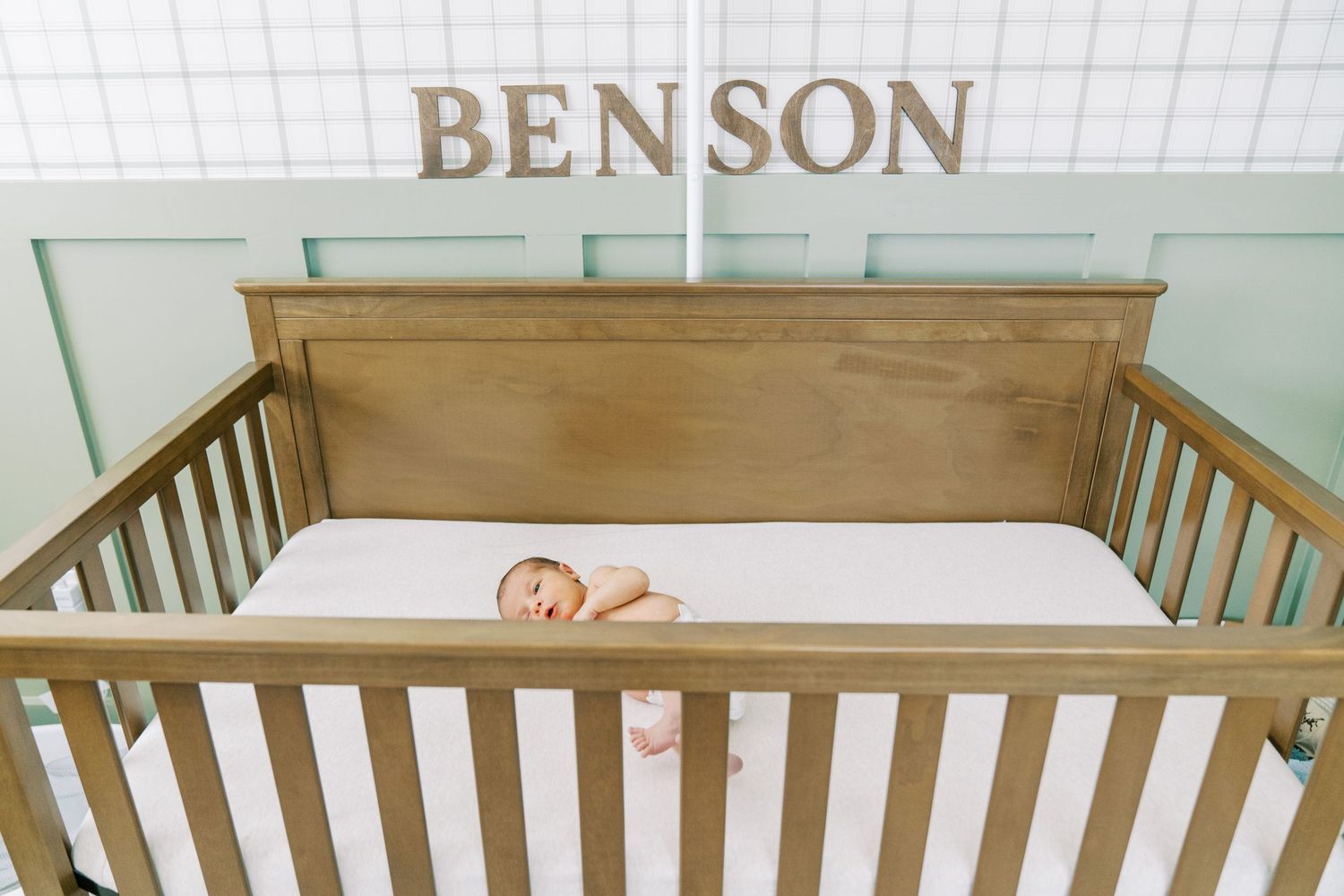 A wooden crib with a white mattress sits against a light green wall in a nursery setting.