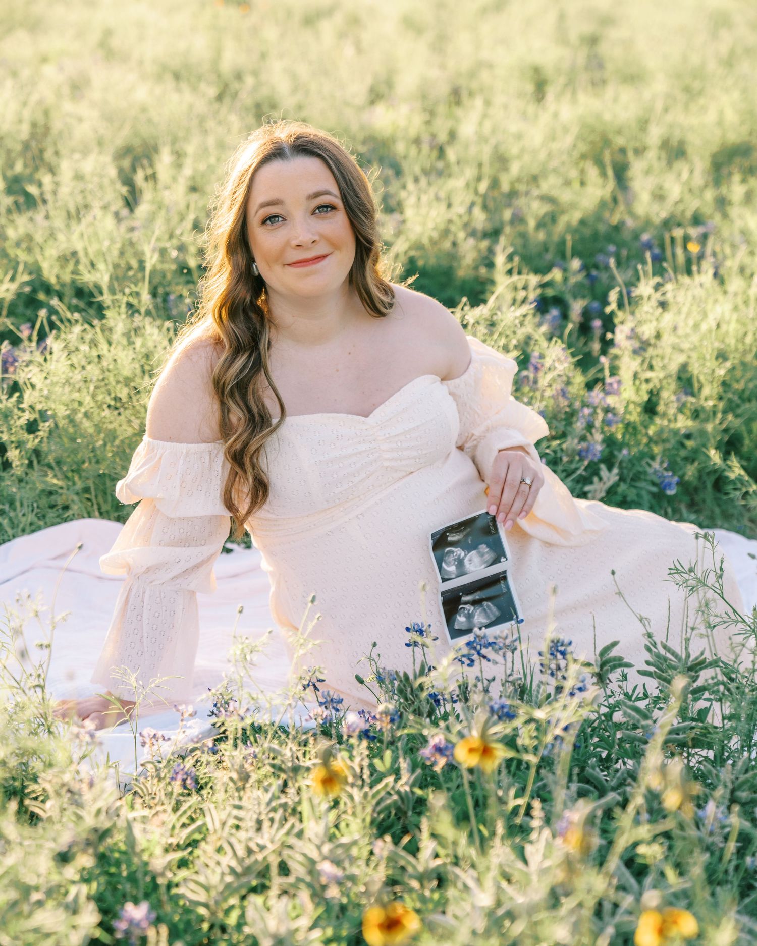 Pregnant woman in blush dress sitting in wildflower field holding ultrasound photos during maternity photoshoot.