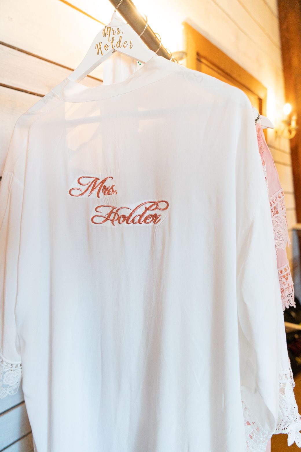White bridal robes with embroidered text hanging on wooden hangers in a rustic setting.