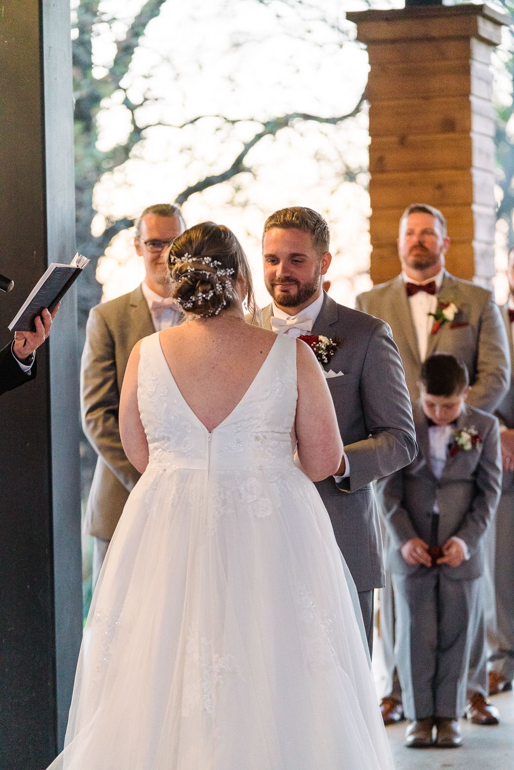 A wedding ceremony takes place on a covered porch with guests in gray suits watching the couple exchange vows.